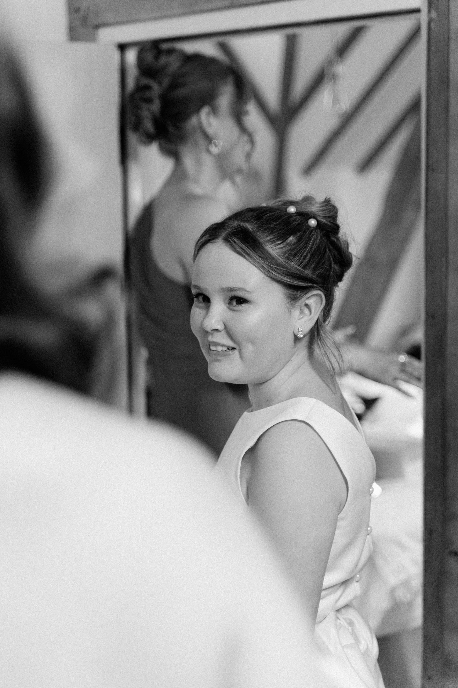 Black and white photo of a smiling bride in a white dress looking over her shoulder, with her reflection visible in a mirror.