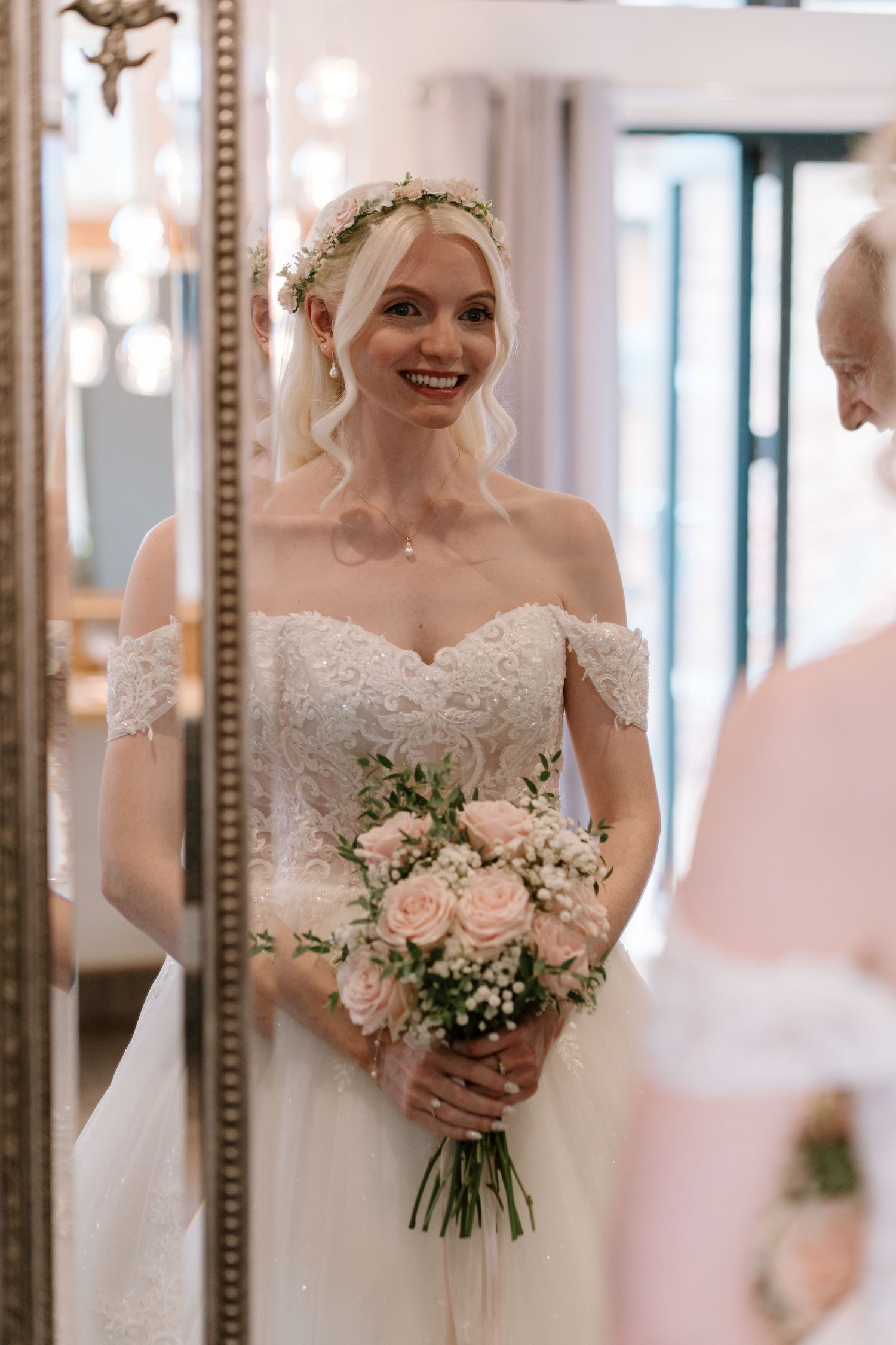 Smiling bride in off-shoulder lace gown holding pink rose bouquet, wearing floral crown, reflected in ornate gold mirror.
