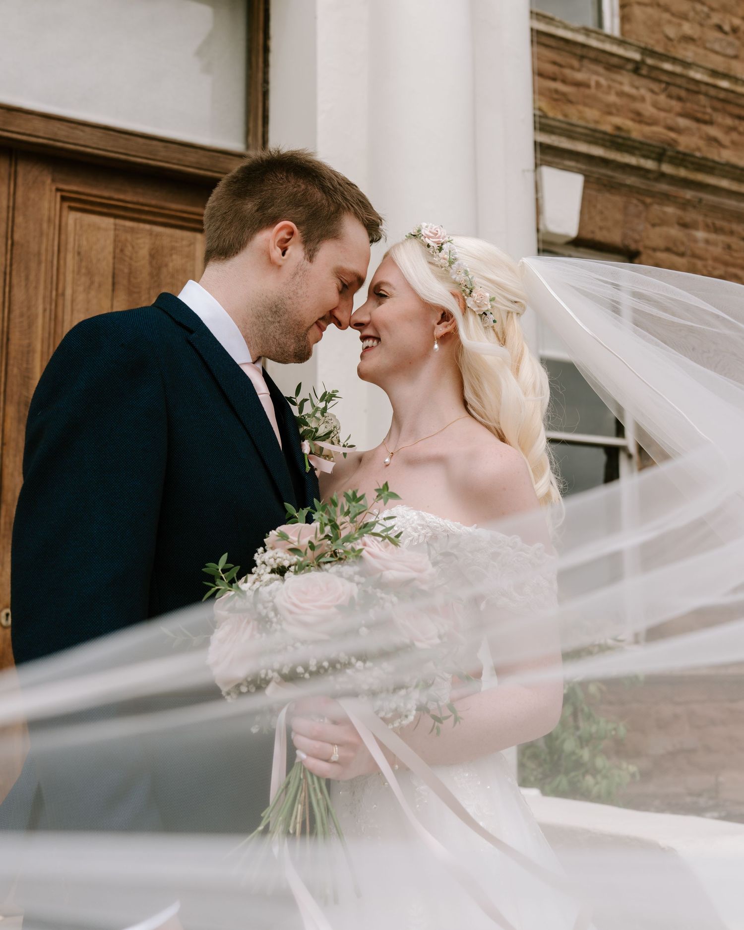 Bride and groom share a romantic forehead touch, veil flowing, holding blush bouquet outside a historic venue.