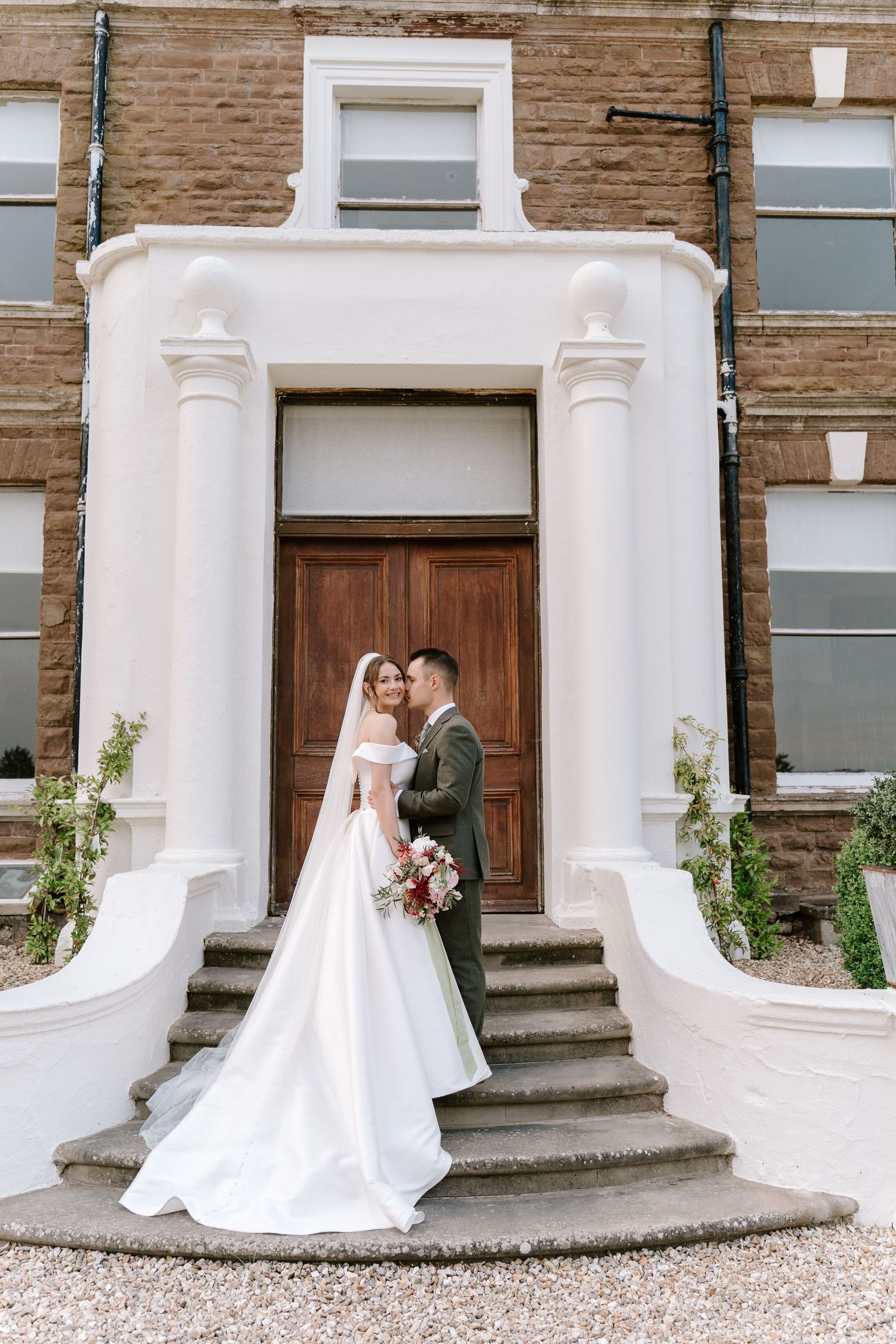 Bride and groom posing on steps of elegant white-columned entrance of brick building, bride holding floral bouquet.
