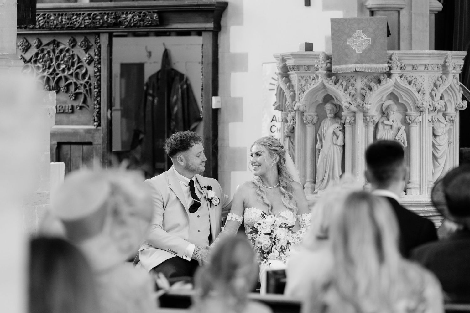 Black and white photo of a smiling bride and groom seated together during their wedding ceremony in a church.