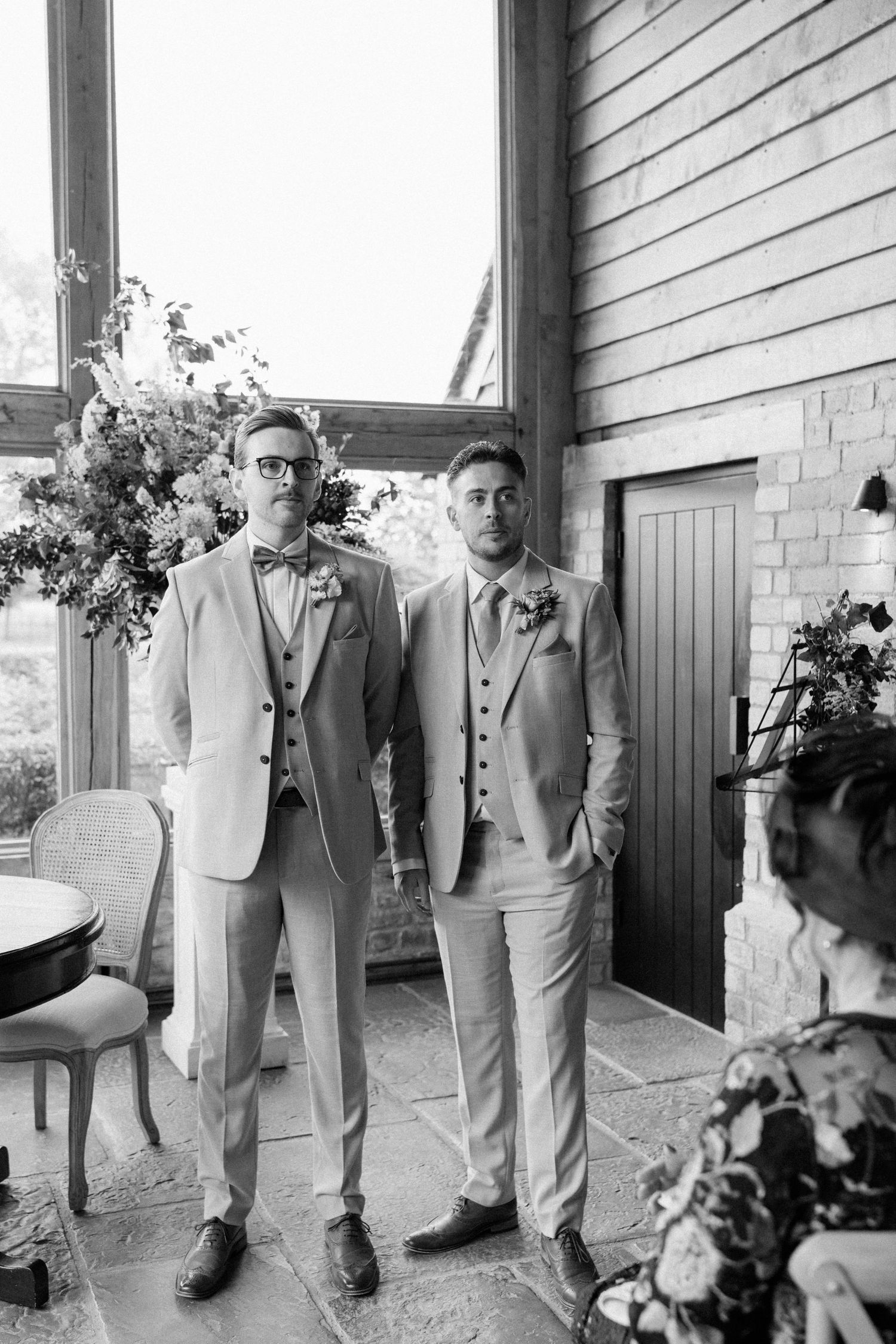 Two grooms in matching light suits with bow ties stand together at their wedding ceremony in an elegant barn venue.