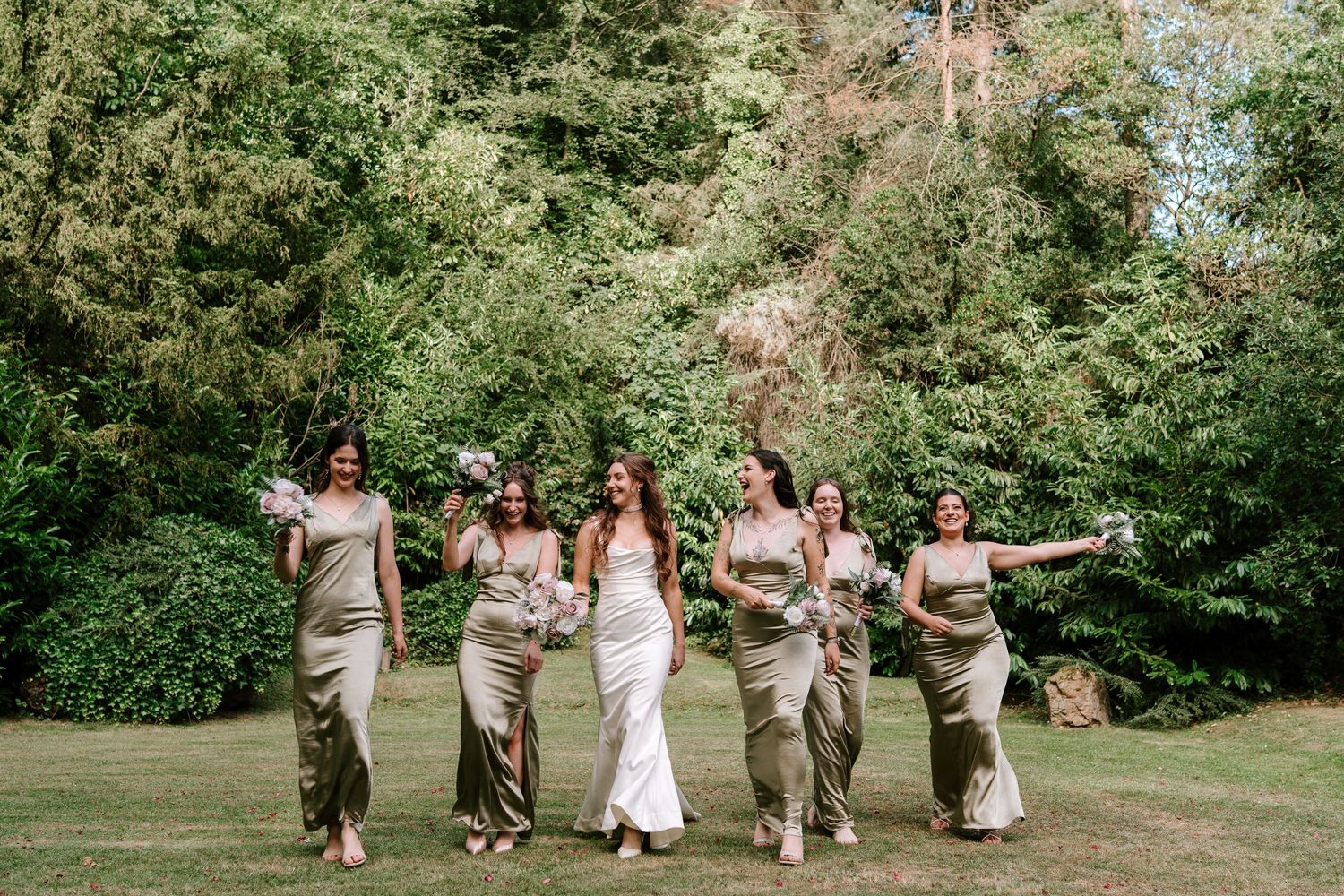 Bride in white gown walks with five bridesmaids in olive satin dresses on green lawn with lush trees.