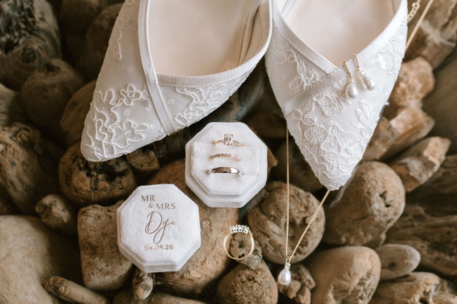 White lace bridal flats displayed with wedding rings in an octagonal ring box on natural stones.