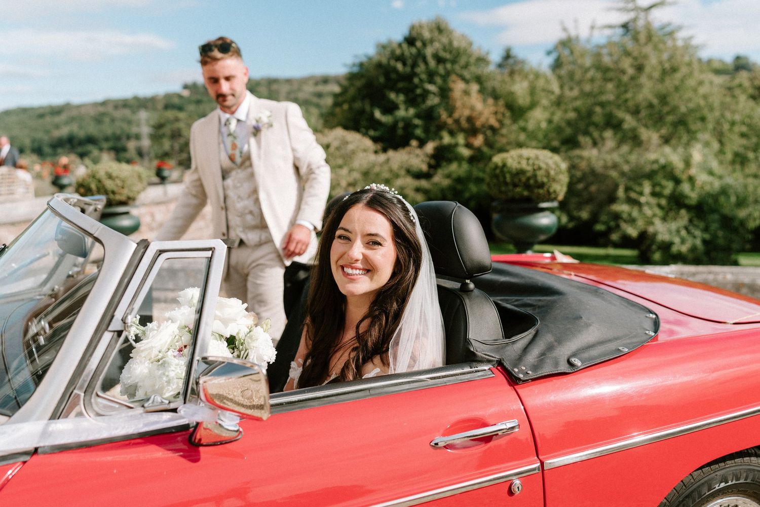 Smiling bride sitting in a red vintage convertible car with groom standing behind her outdoors.