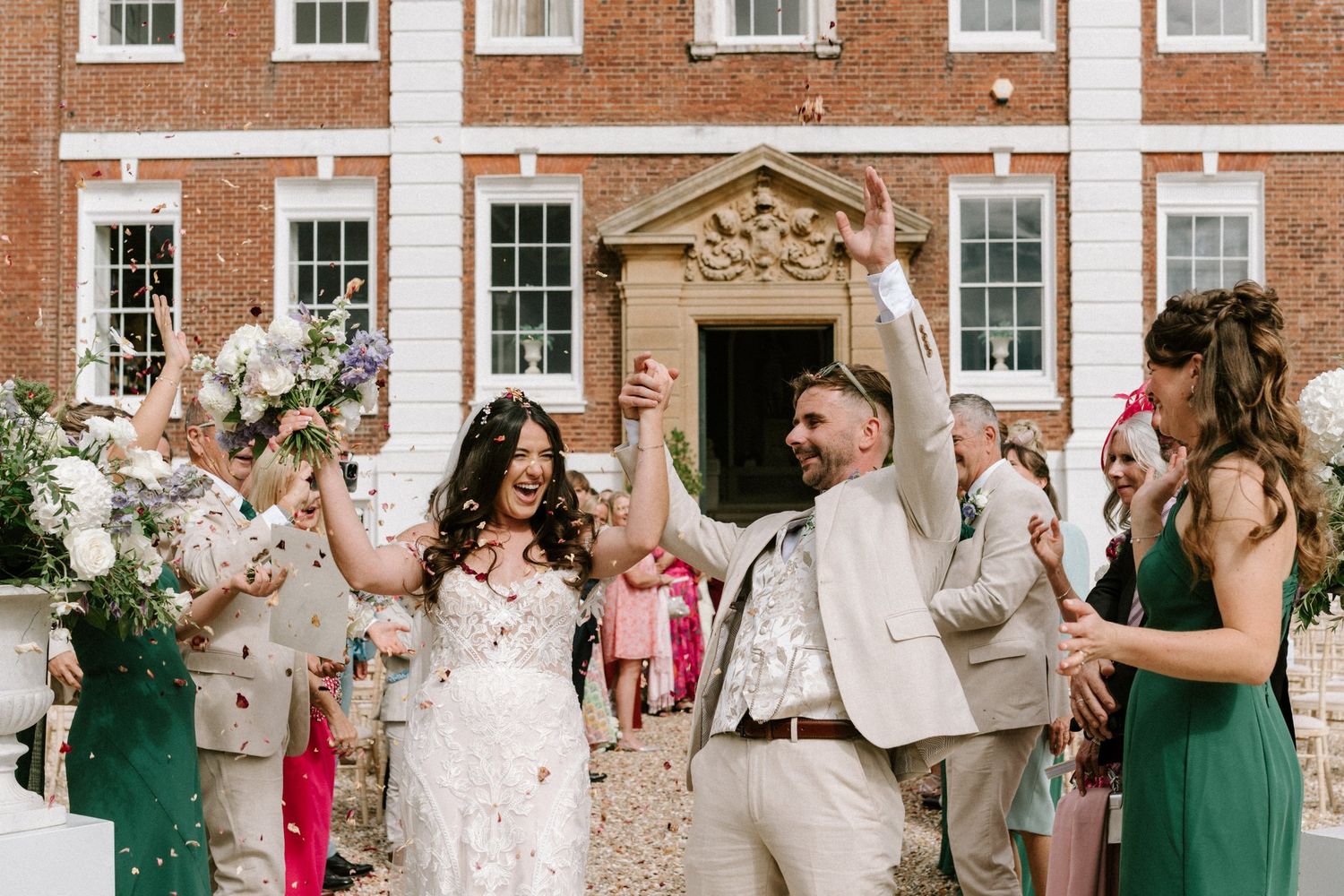 Joyful newlyweds exit wedding venue amid confetti shower, bride in lace gown, groom in tan suit, guests celebrating.
