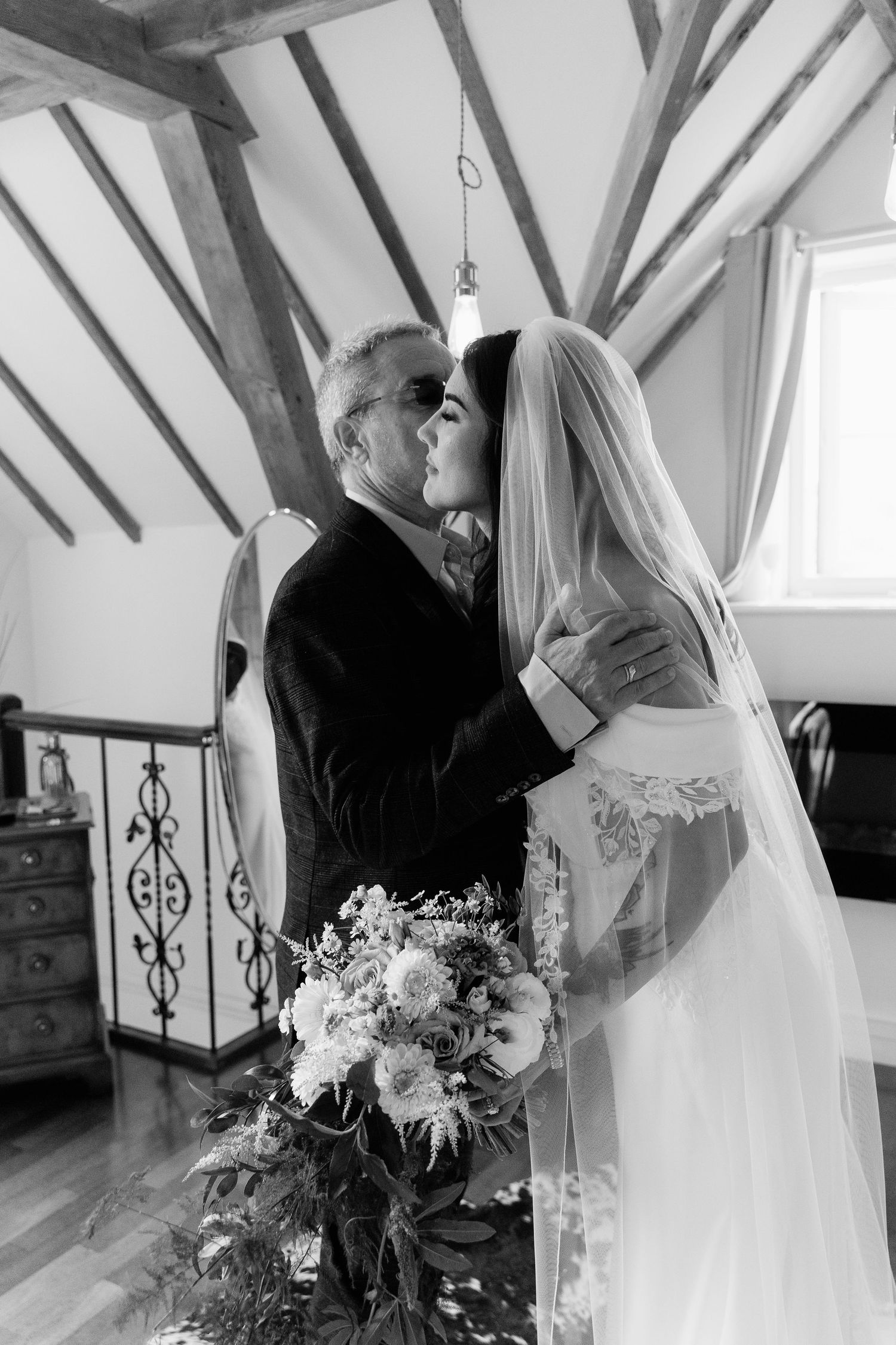 Black and white photo of a bride in a veil kissing an older man, holding a floral bouquet in a rustic venue.