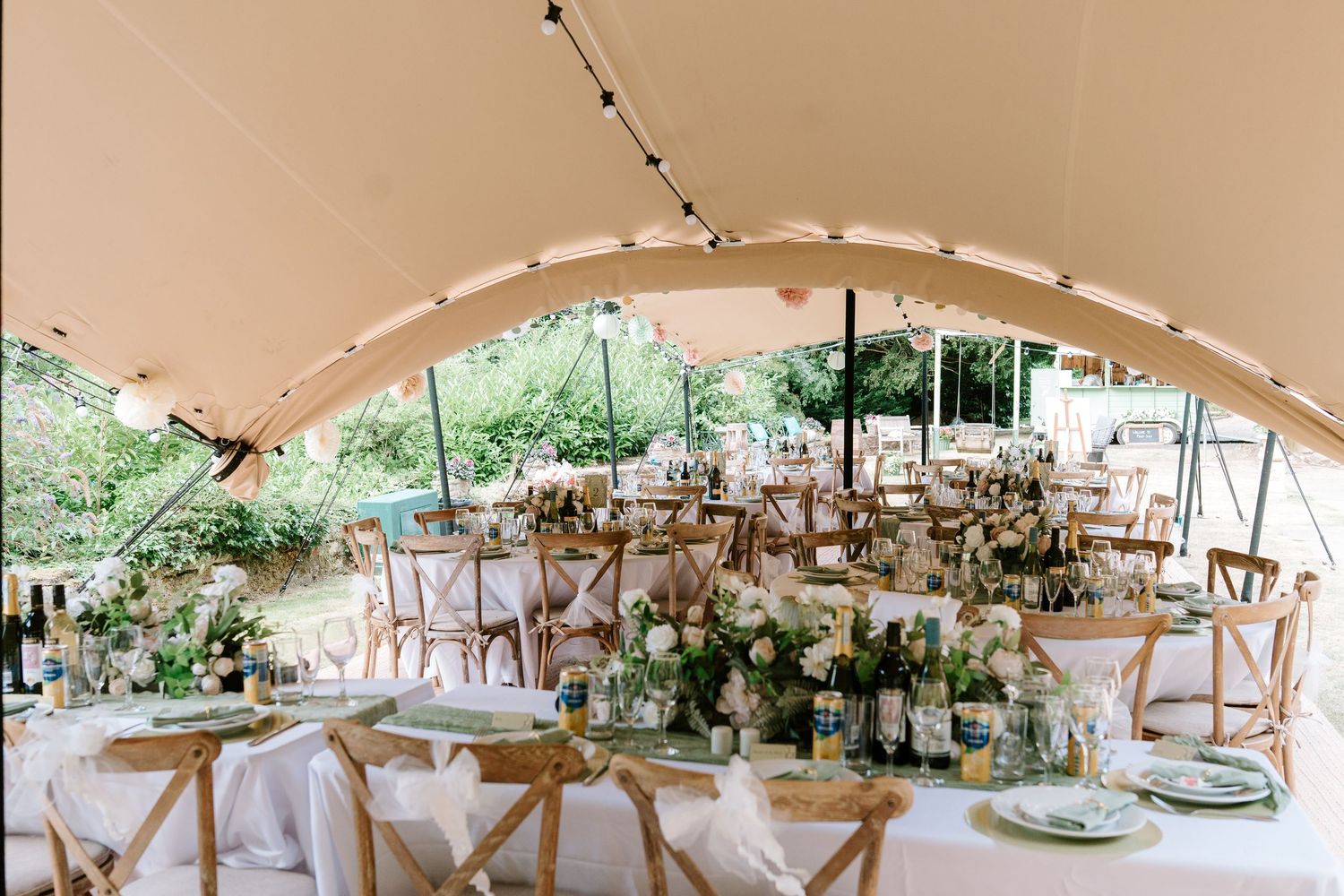 Elegant outdoor wedding reception under a marquee tent with floral centerpieces, string lights, and wooden cross-back chairs.