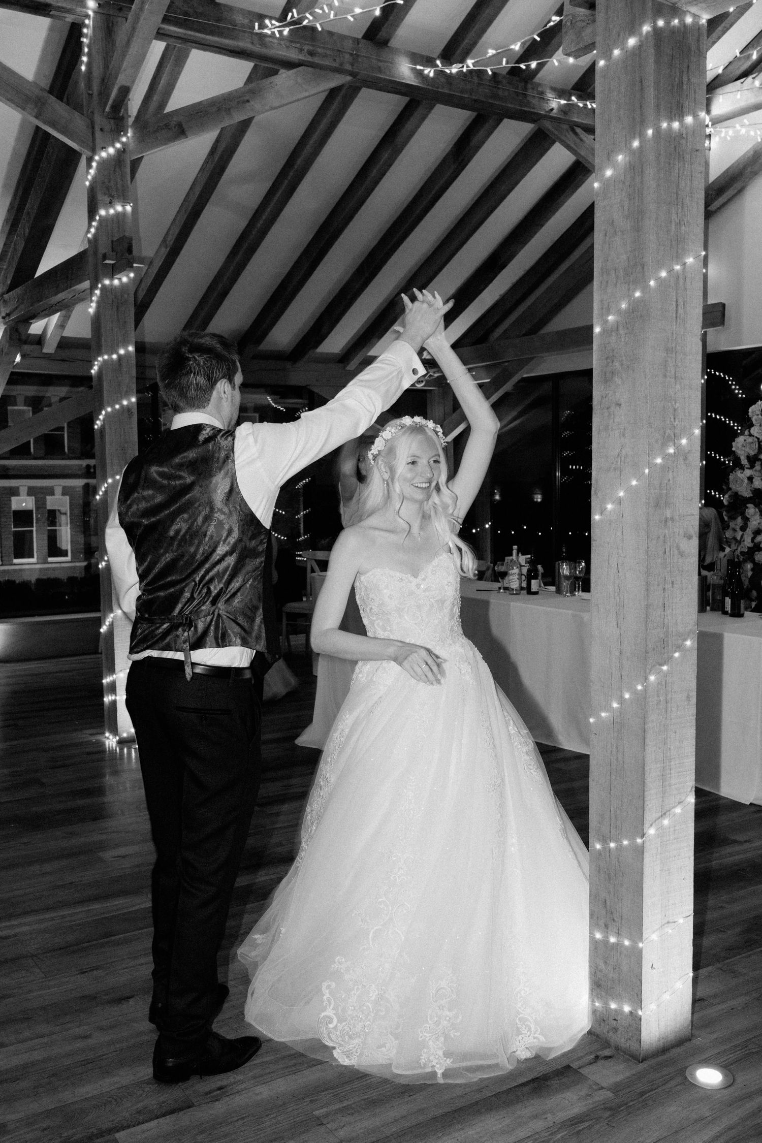 Bride and groom share their first dance, twirling under fairy lights in a rustic barn venue, captured in black and white.