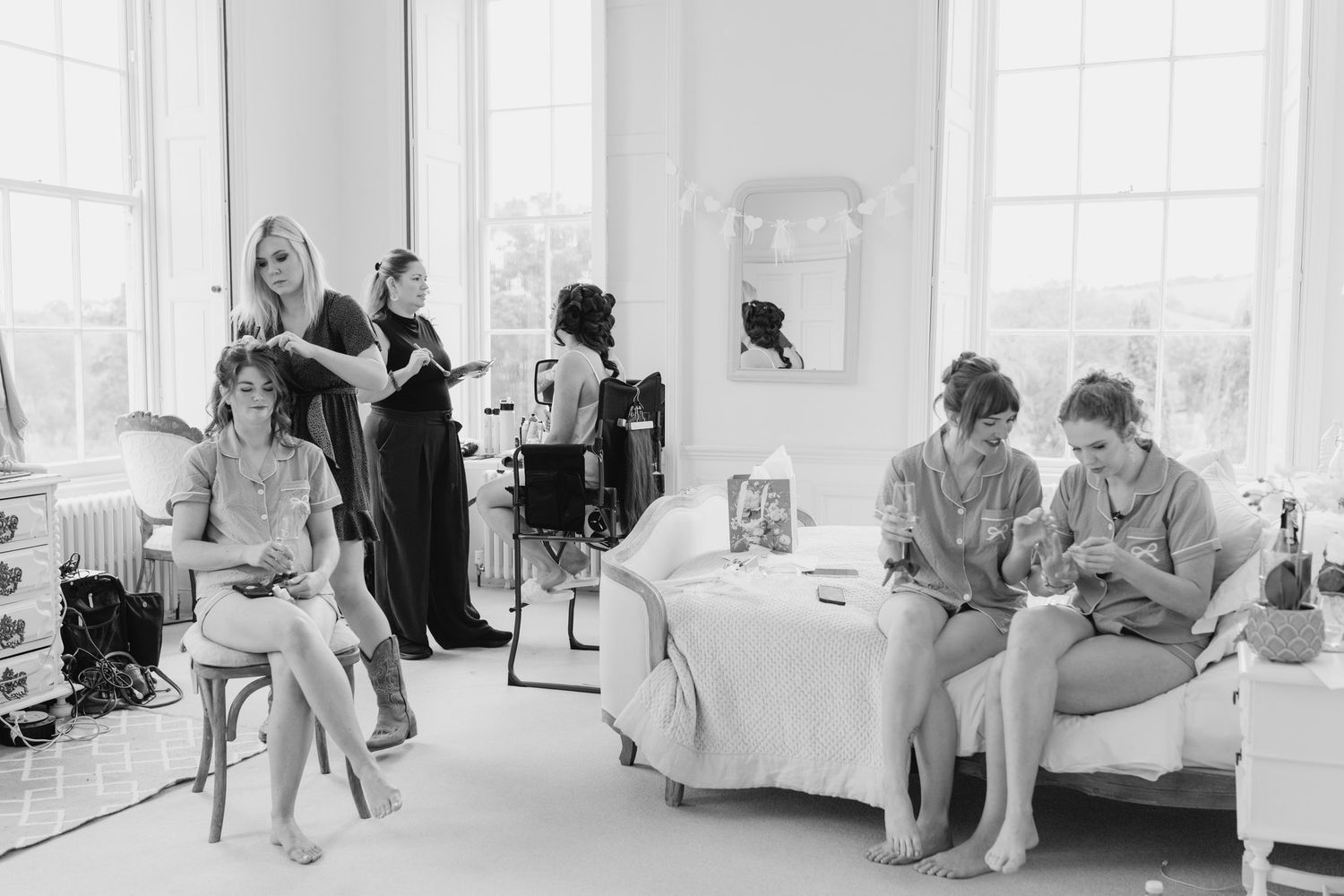 Black and white photo of bridesmaids getting ready, with hair styling and relaxed conversation in a bright, elegant room.