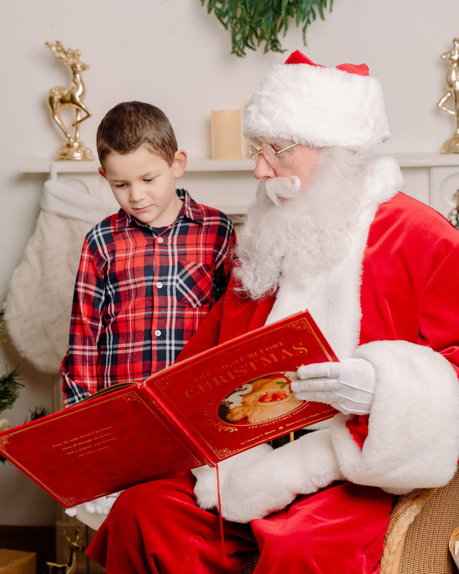 A young boy in plaid pajamas reads a Christmas book with Santa Claus beside a decorated fireplace.
