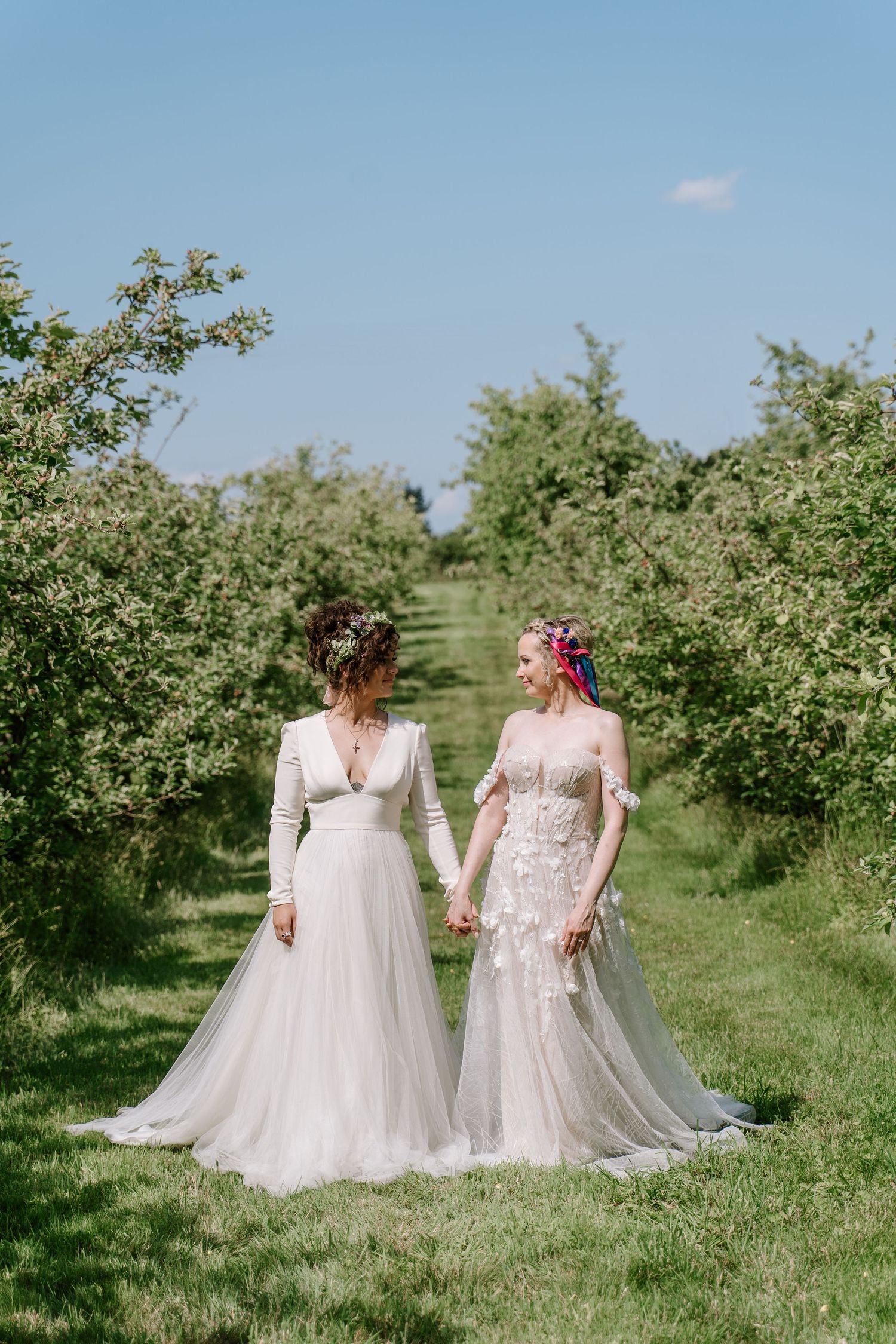 Two brides in white wedding gowns hold hands walking through a lush green orchard on a sunny day.