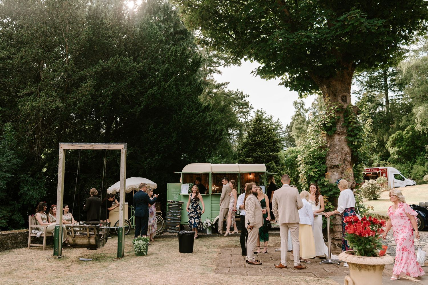 Wedding guests mingling outdoors near vintage green catering truck surrounded by lush trees.