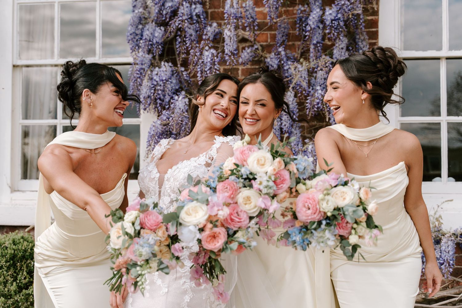 Bride and two bridesmaids in ivory gowns laughing outdoors, holding pastel floral bouquets with wisteria backdrop.