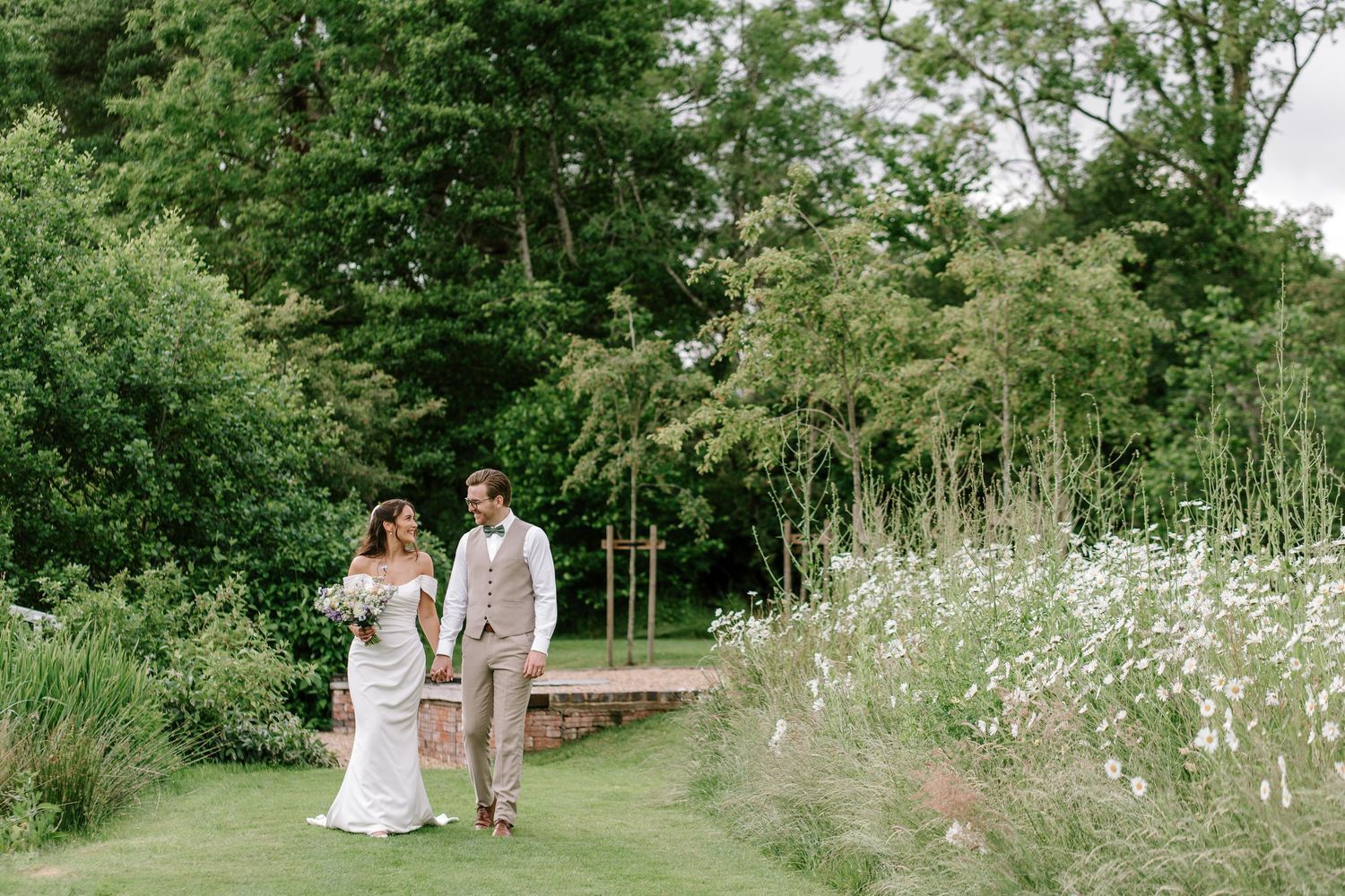 Bride and groom walking hand in hand through lush green garden with wildflowers on their wedding day.