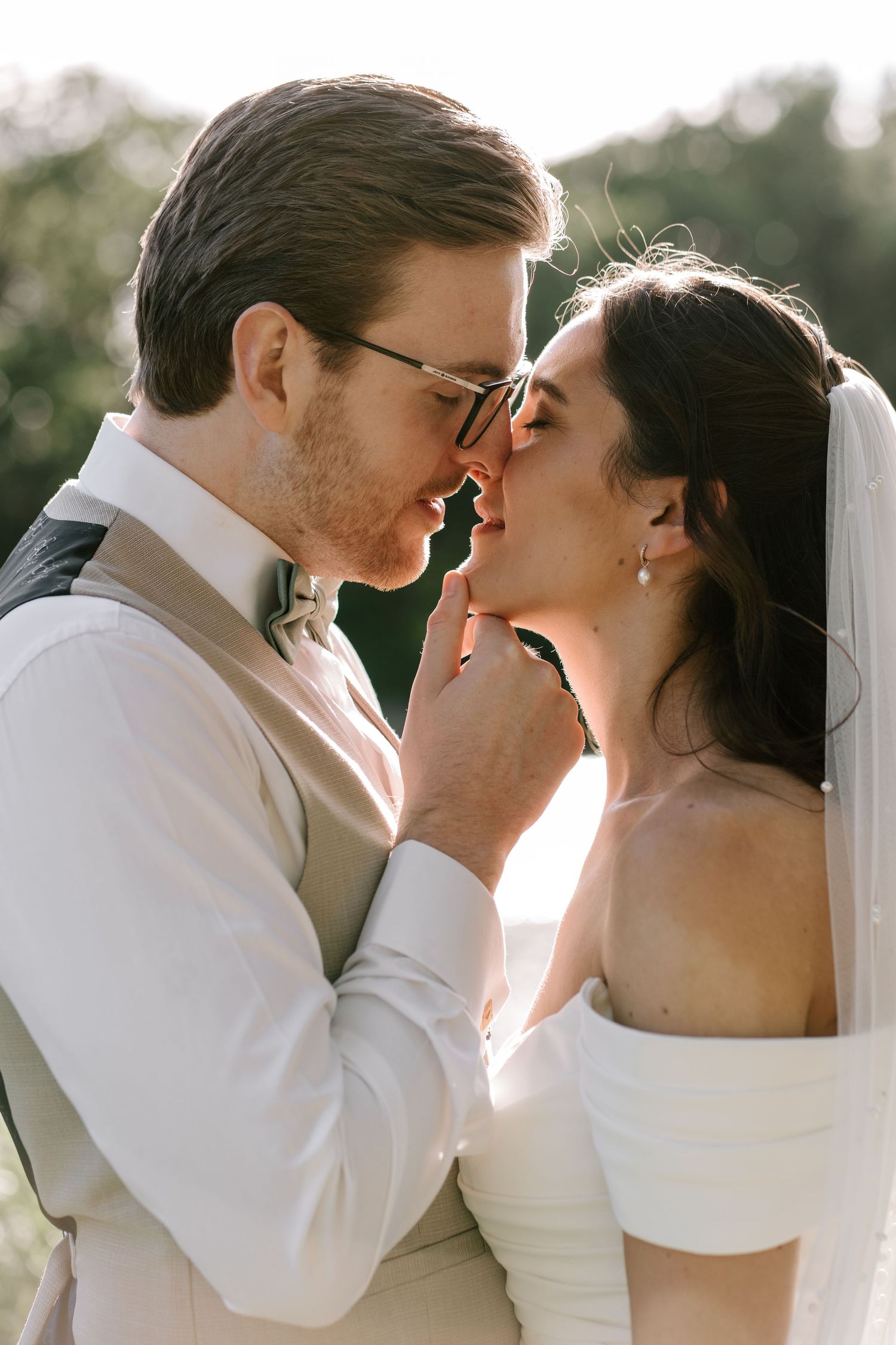 Groom in beige suit tenderly holds brides face as they share a close romantic moment in golden light.