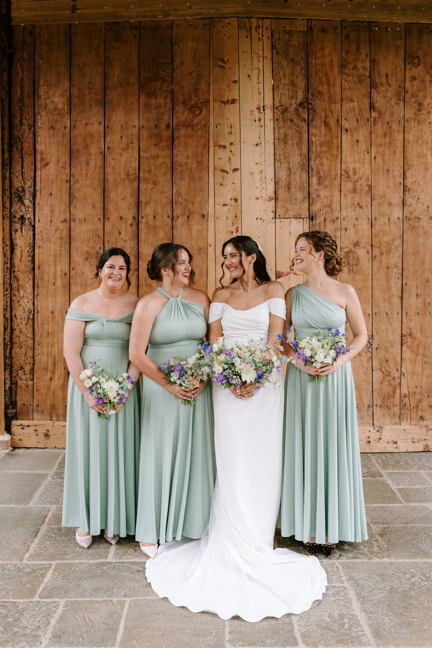 Bride in white gown with three bridesmaids in sage green multiway dresses holding wildflower bouquets before a wooden barn door.