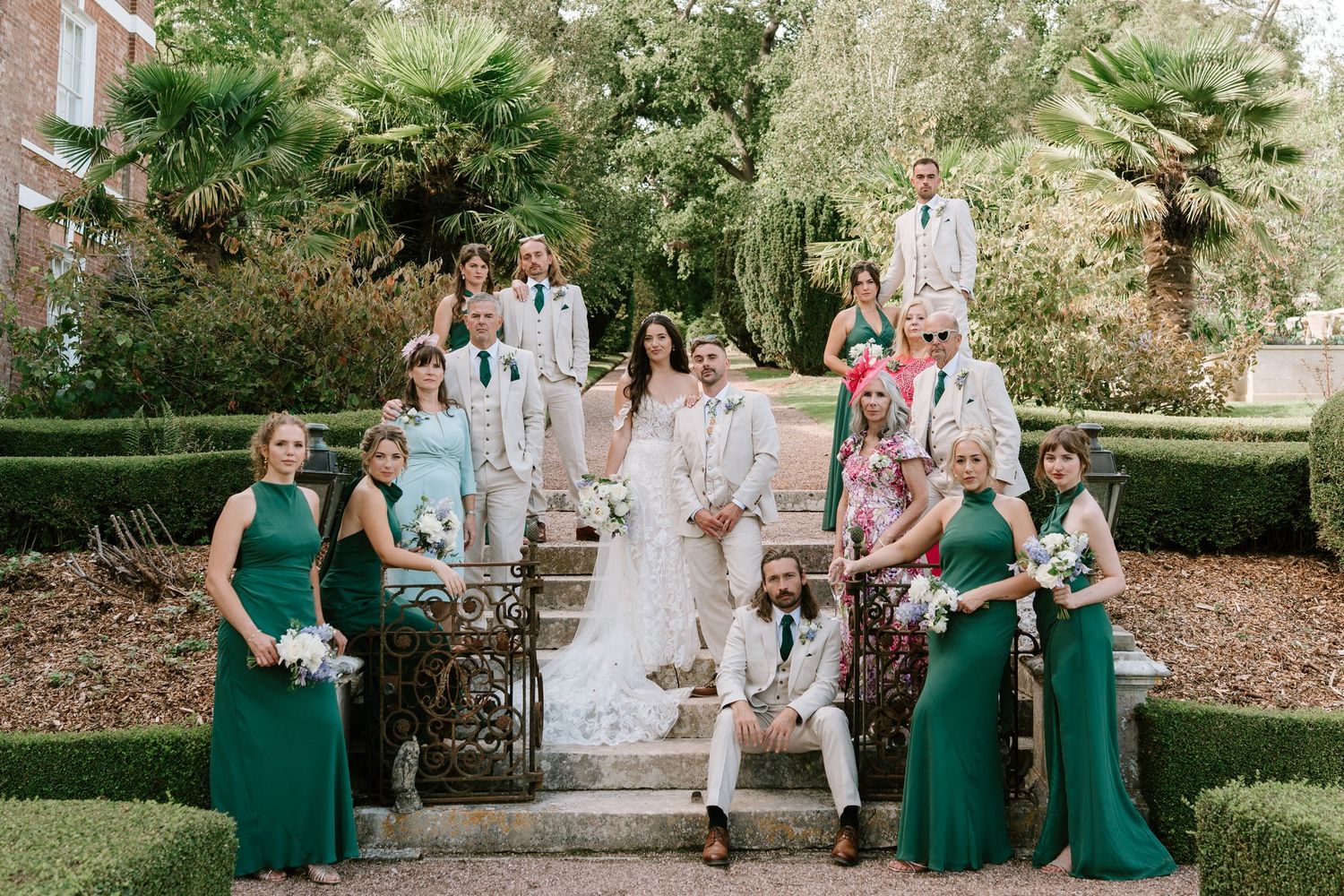 Wedding party poses on garden steps, bridesmaids in emerald green gowns with bride in white lace and groom in ivory suit.