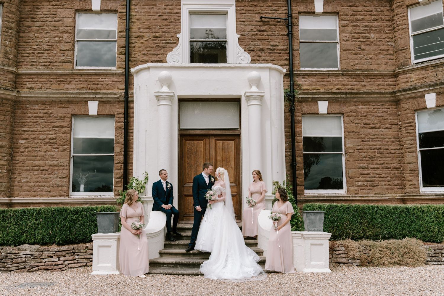 Bride and groom with bridesmaids in pink dresses posing outside a brick manor house wedding venue.