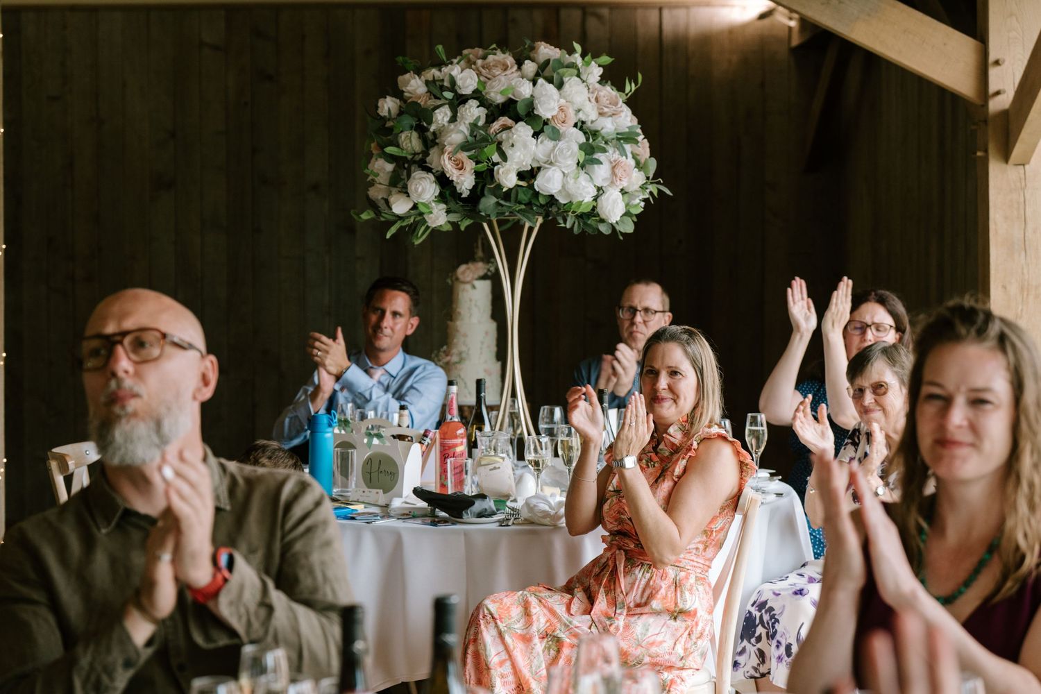 Wedding guests clapping and smiling at a reception, seated at tables with floral centerpieces in a rustic venue.