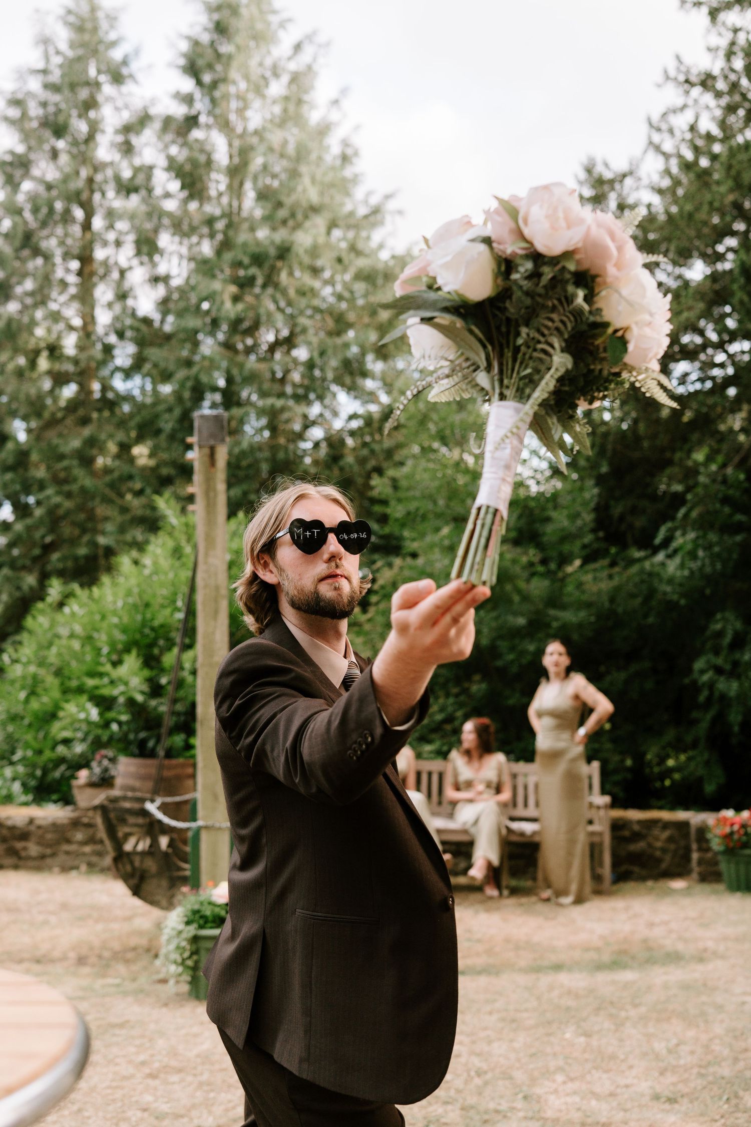 Stylish wedding guest in sunglasses holding bridal bouquet aloft at outdoor garden wedding reception.