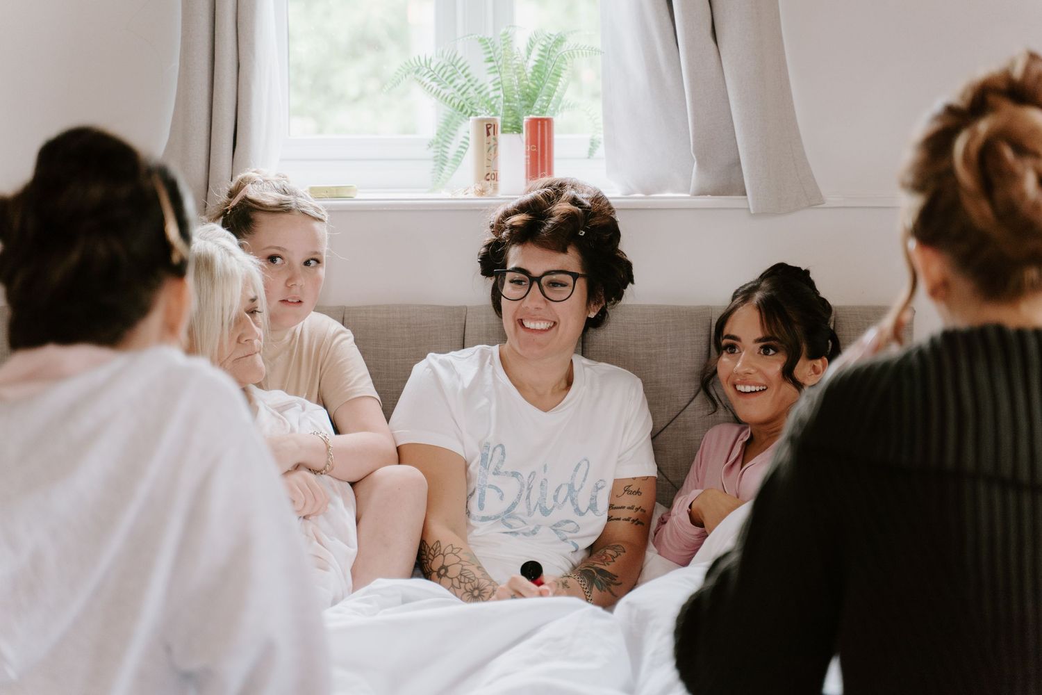 Bride laughing with bridesmaids on a couch during wedding morning preparations in a bright cozy room.
