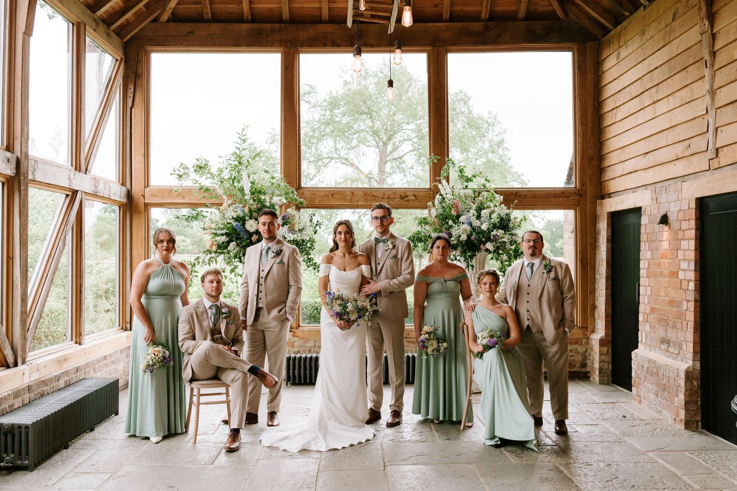 Wedding party posing in a rustic barn venue with bride, groom, groomsmen in tan suits and bridesmaids in sage green dresses.
