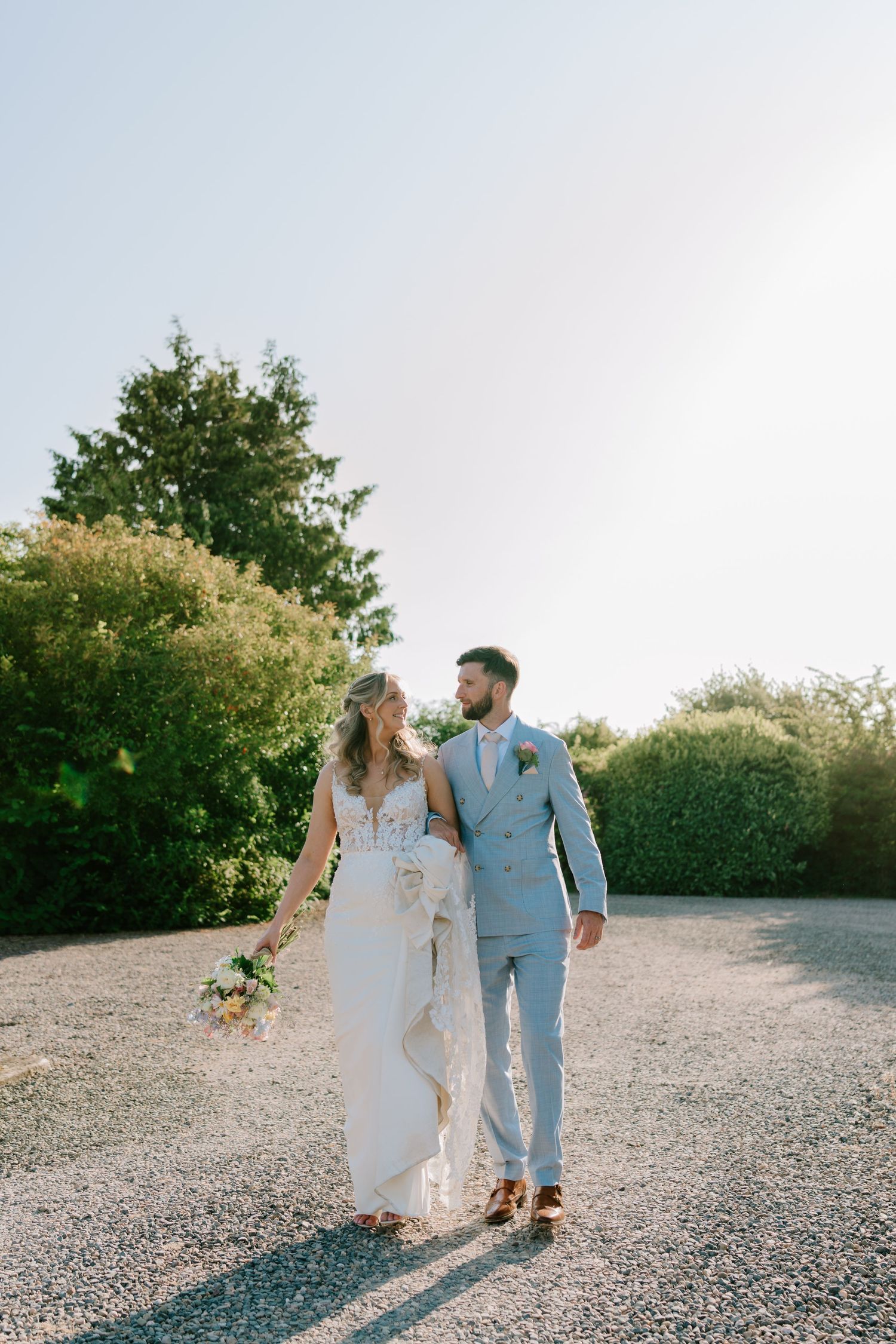 Bride and groom walking hand in hand on a gravel path surrounded by lush greenery on their wedding day.