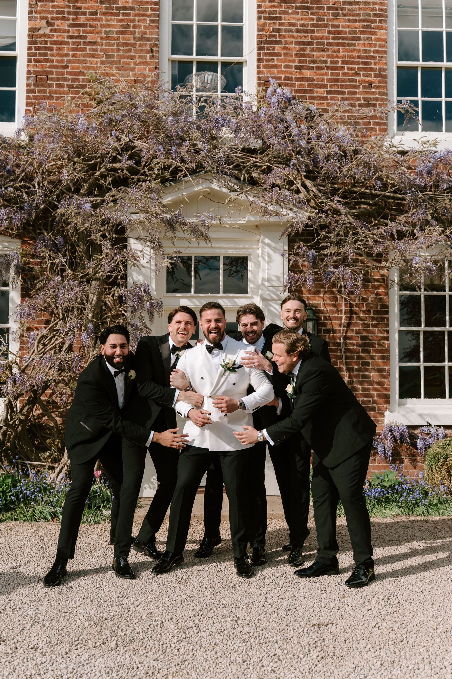 Groom in white tuxedo jacket celebrates with groomsmen in black suits outside brick manor with wisteria.