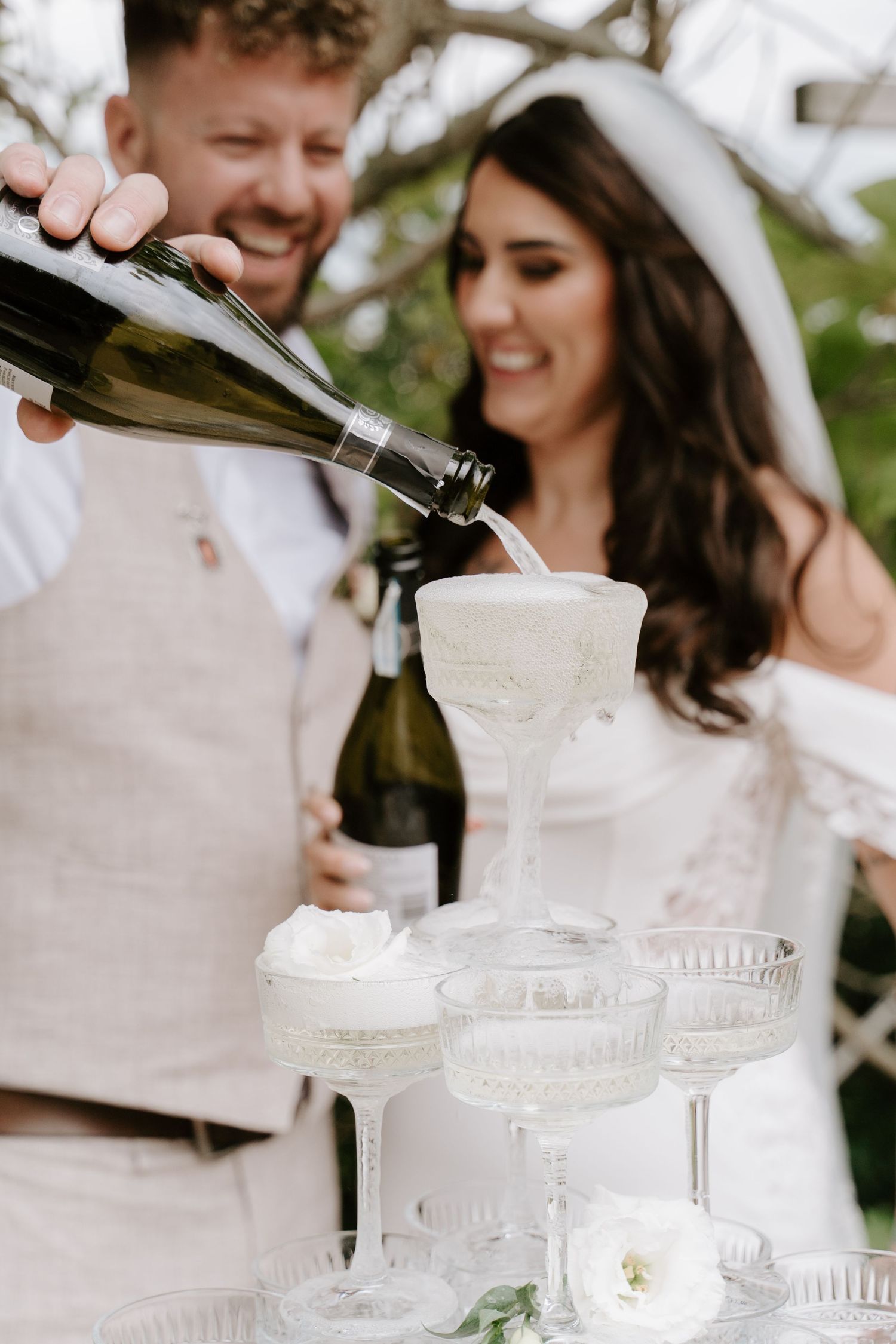 Bride and groom pouring champagne tower at wedding reception outdoors.