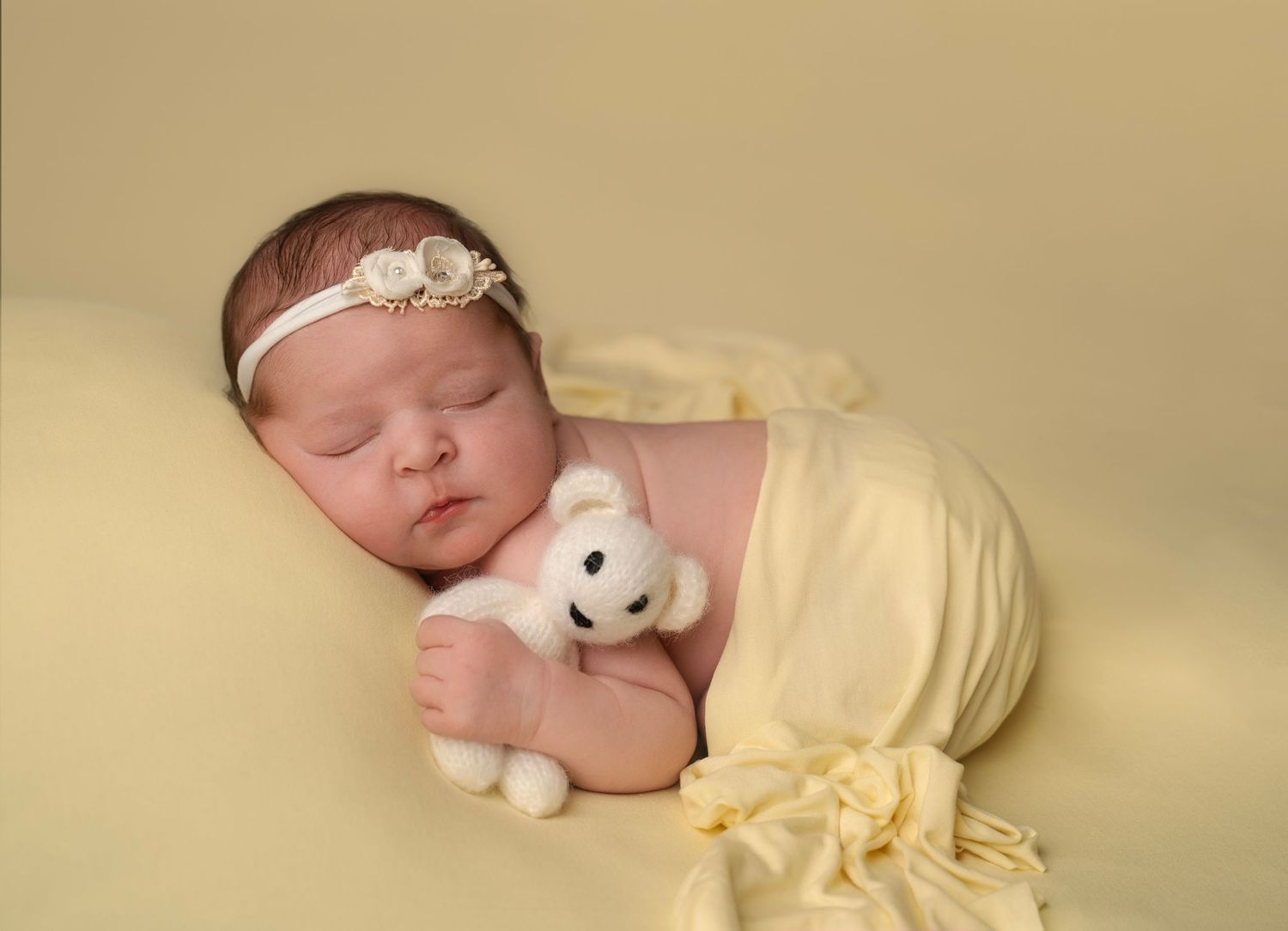 Sleeping newborn baby wrapped in yellow cloth holding a white teddy bear with a floral headband.