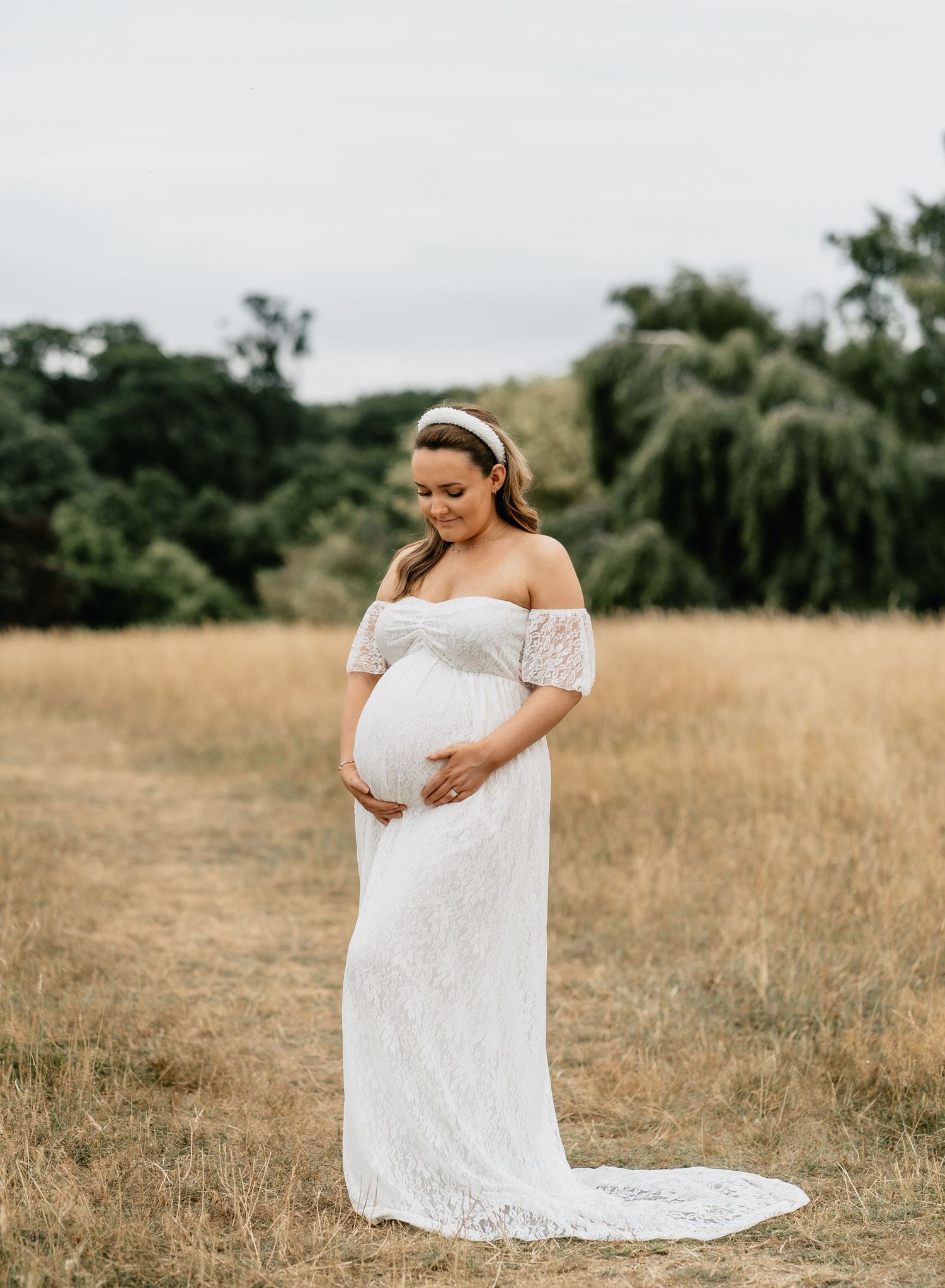 Pregnant woman in white off-shoulder lace gown holding baby bump in golden field during outdoor maternity photoshoot.