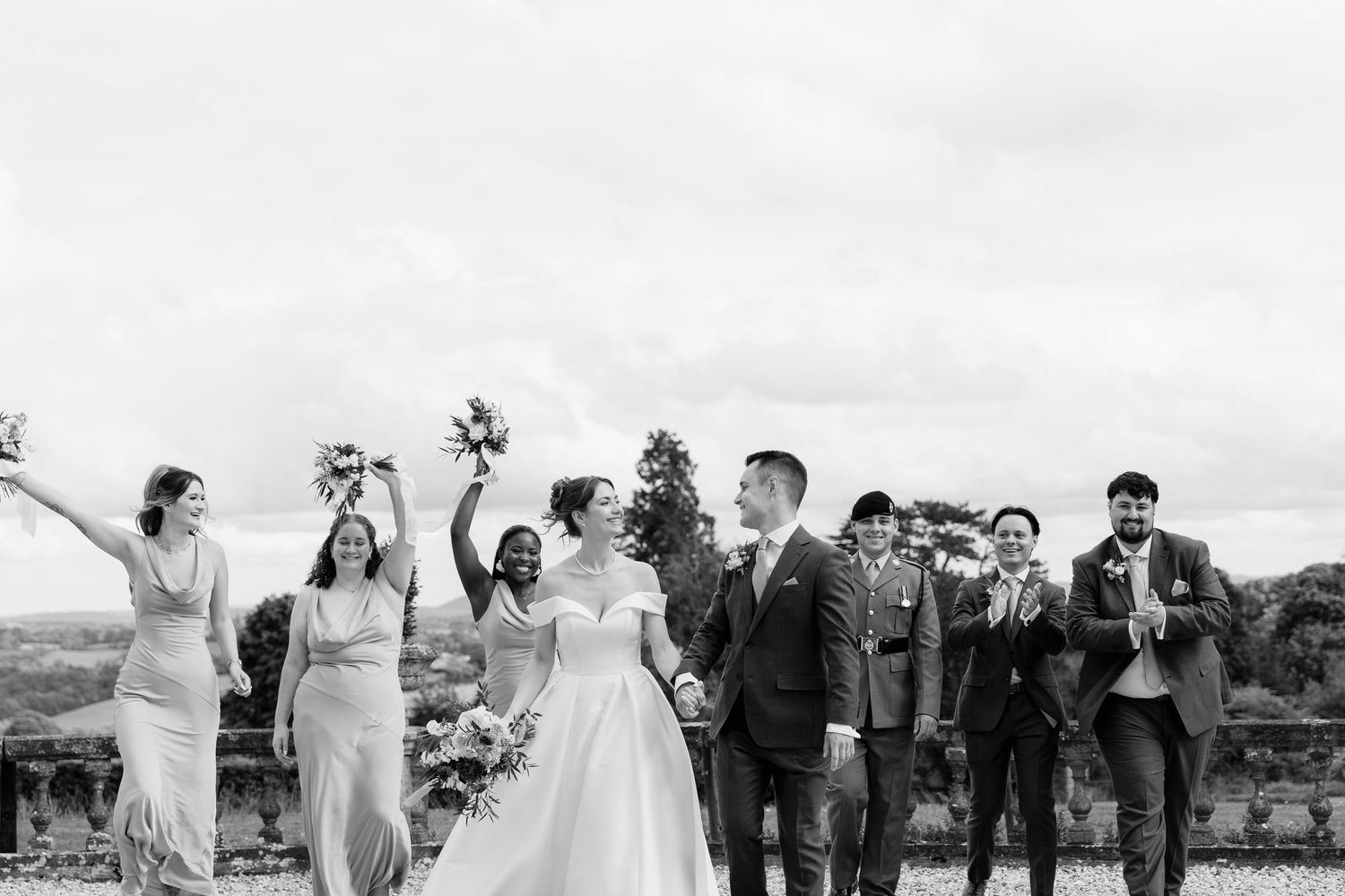 Black and white wedding photo of bride and groom celebrating with bridal party outdoors at scenic venue.