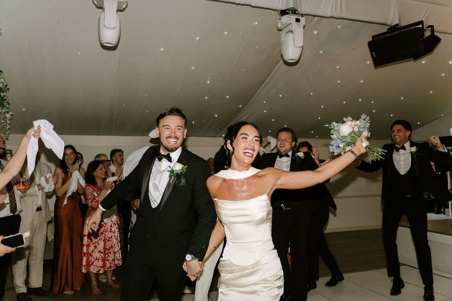 Joyful newlyweds enter wedding reception holding hands, bride in white gown with bouquet, groom in black tuxedo.