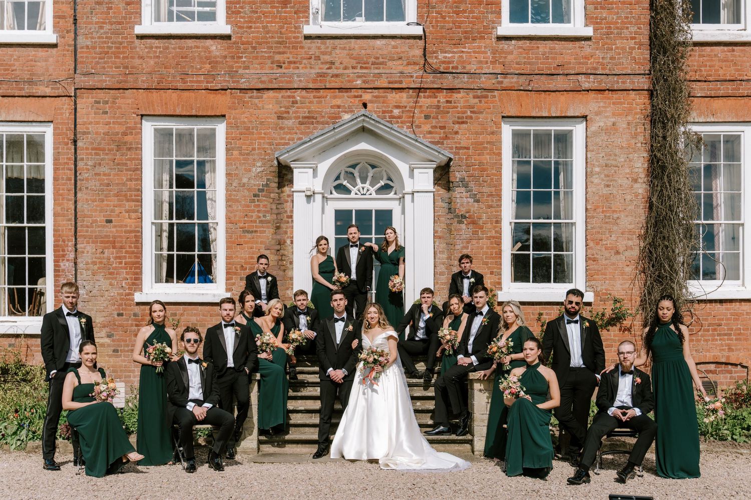 Wedding party in emerald green gowns and black tuxedos posed on steps of elegant red brick manor house.