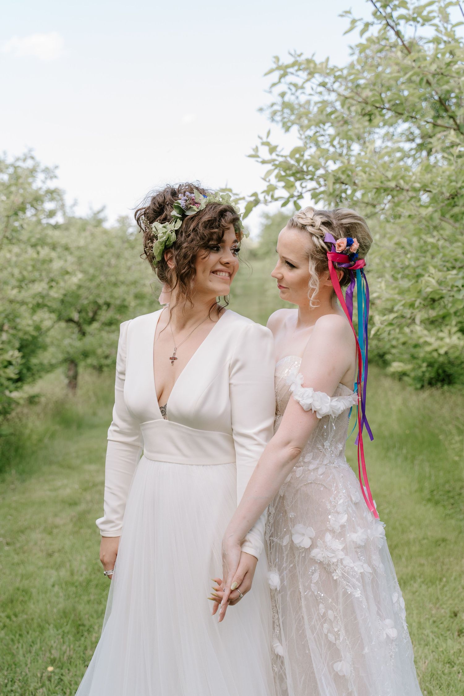 Two brides in white wedding dresses with floral crowns hold hands outdoors among blooming trees.