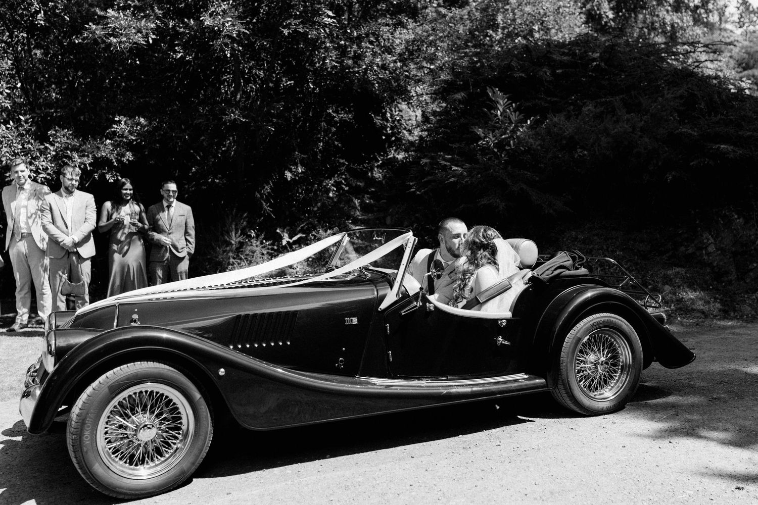 Vintage black and white photo of couple riding in classic open-top sports car on tree-lined road with onlookers.
