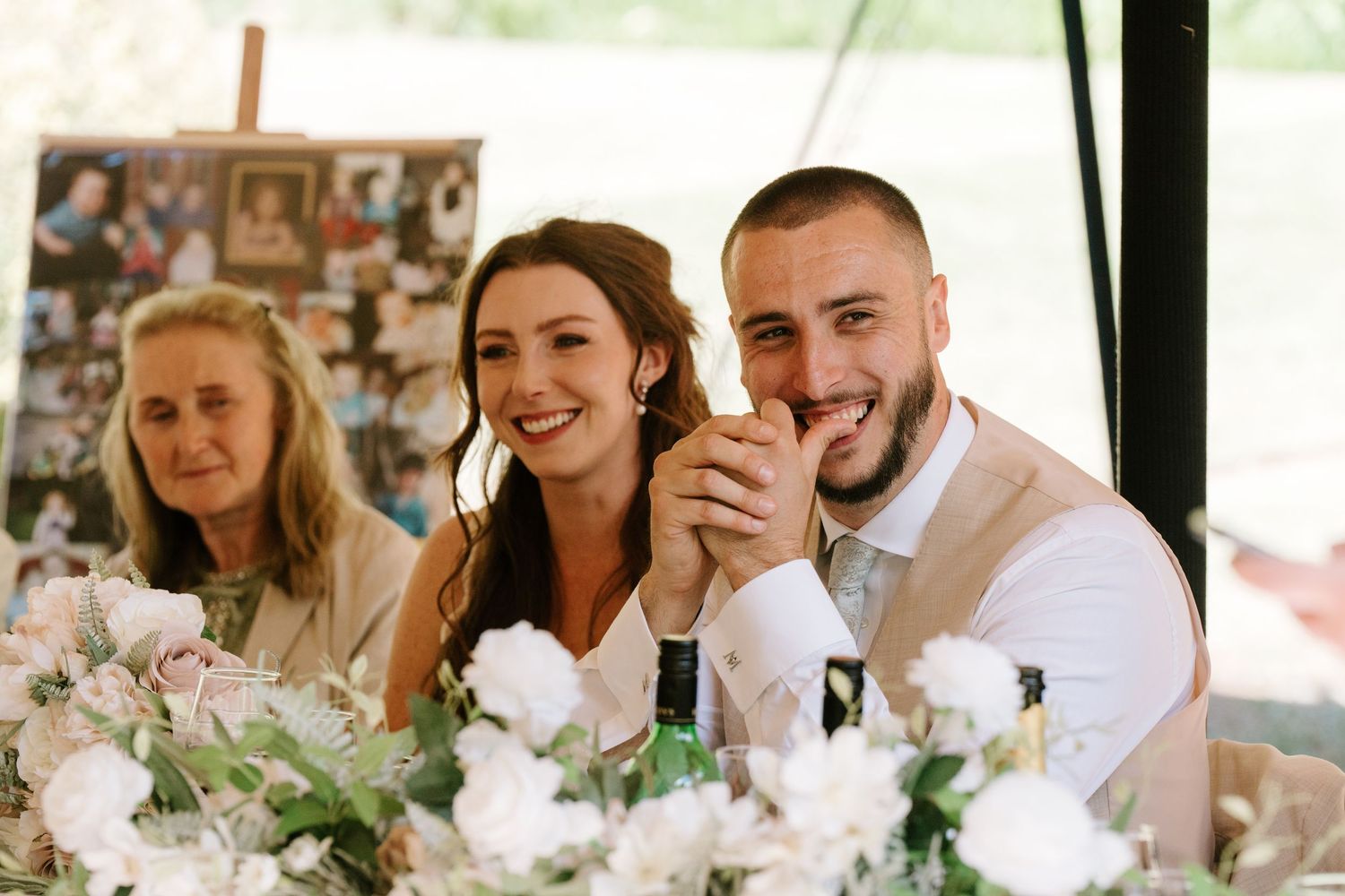 Bride and groom smiling warmly at head table with white floral centerpieces during wedding reception.