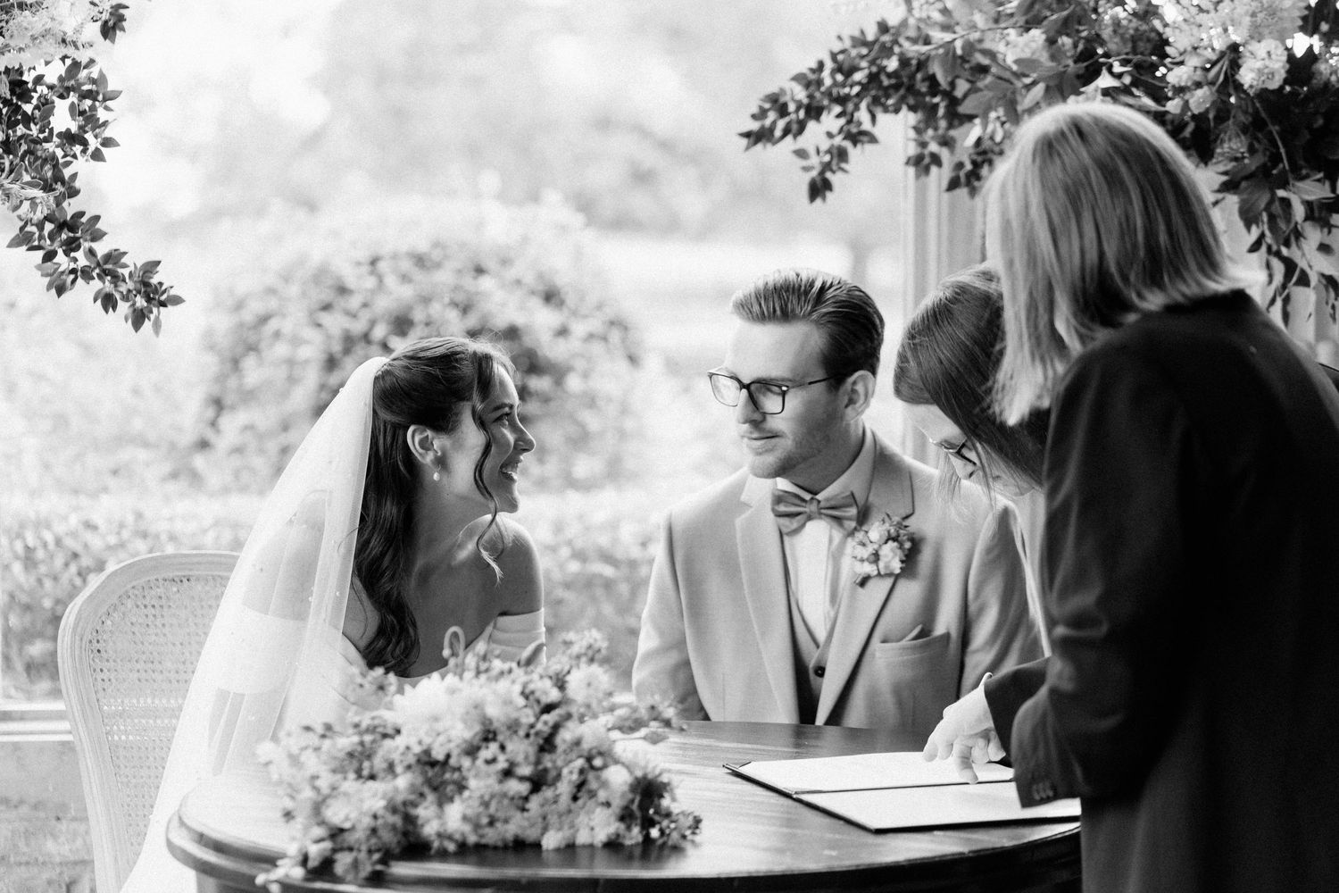 Bride and groom signing their wedding register at an outdoor ceremony table adorned with floral arrangements.