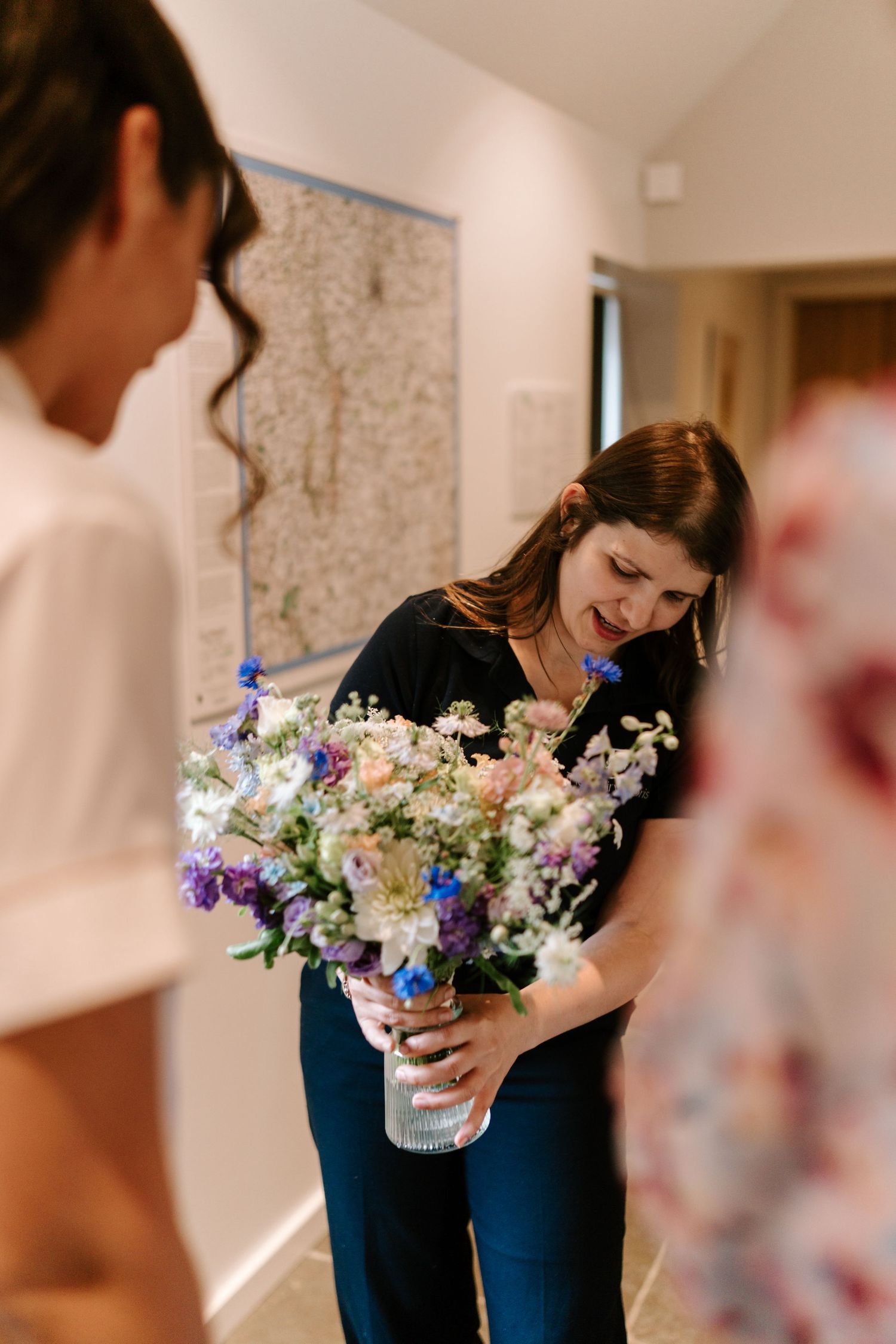 Woman in navy outfit smiling while holding colorful wildflower bouquet in vase indoors.