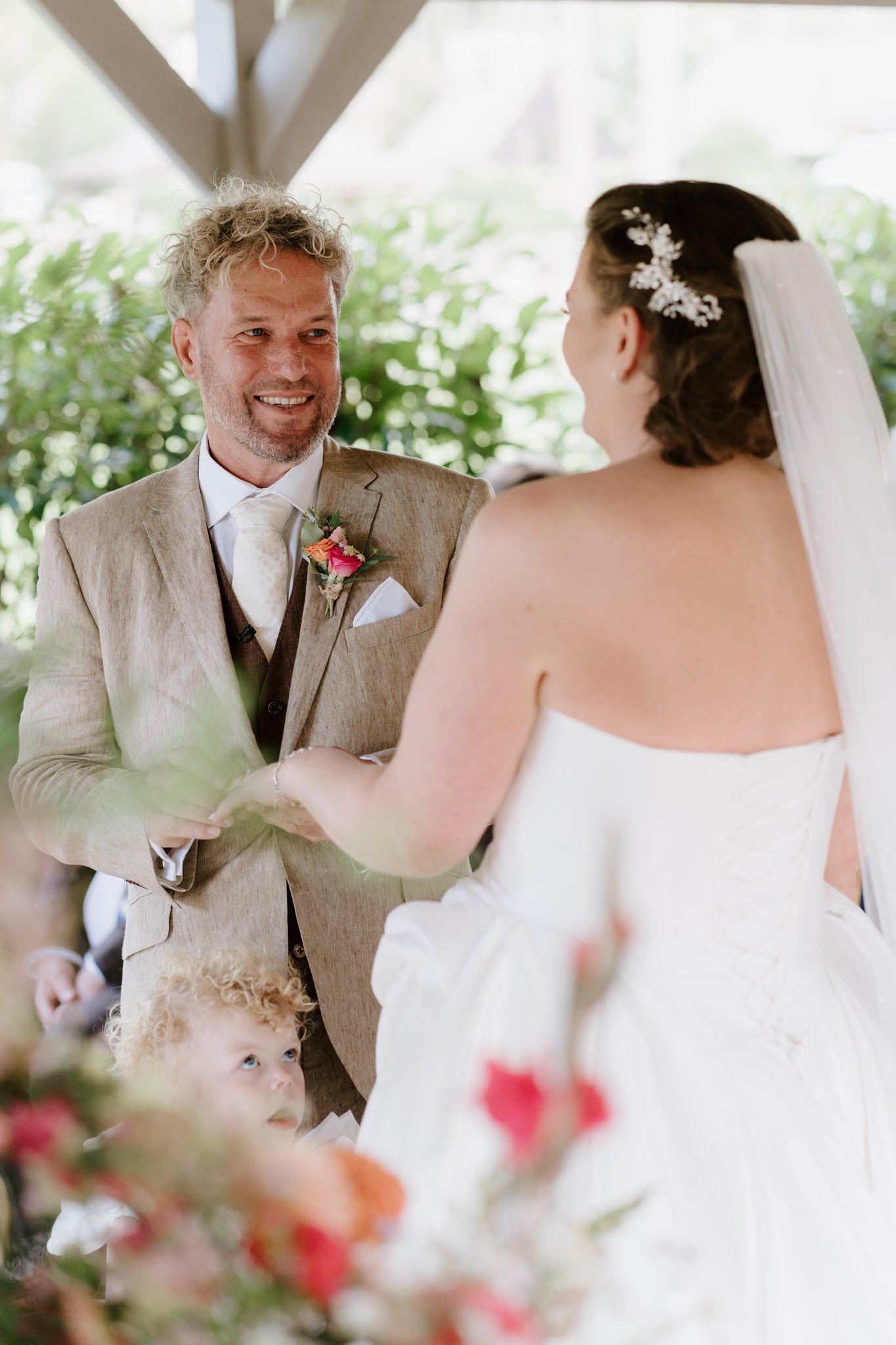 Groom in linen suit smiles at bride during outdoor wedding ceremony while a young child stands nearby.