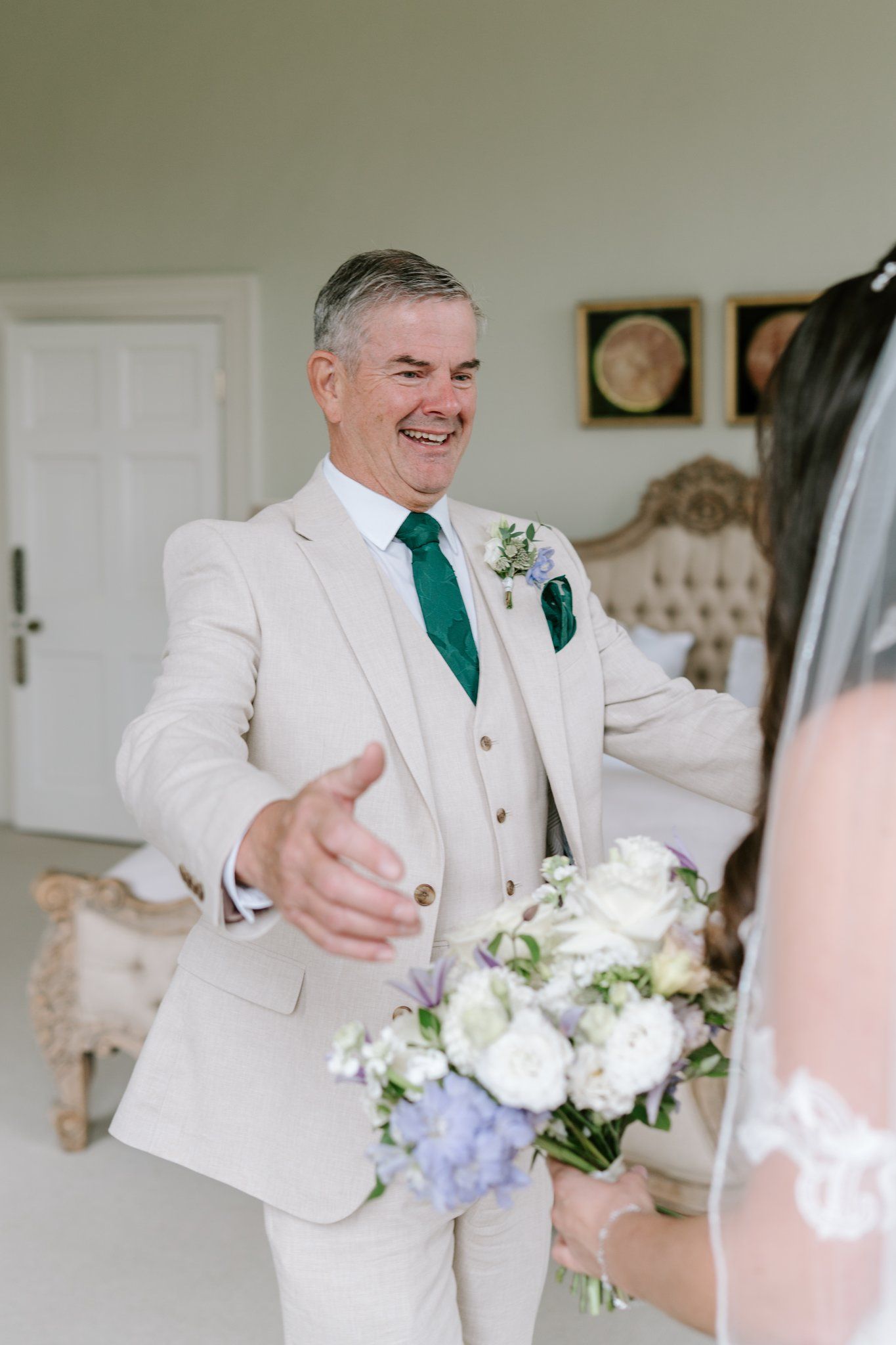Smiling groom in beige suit with green tie reaches out to bride holding white and blue floral bouquet indoors.