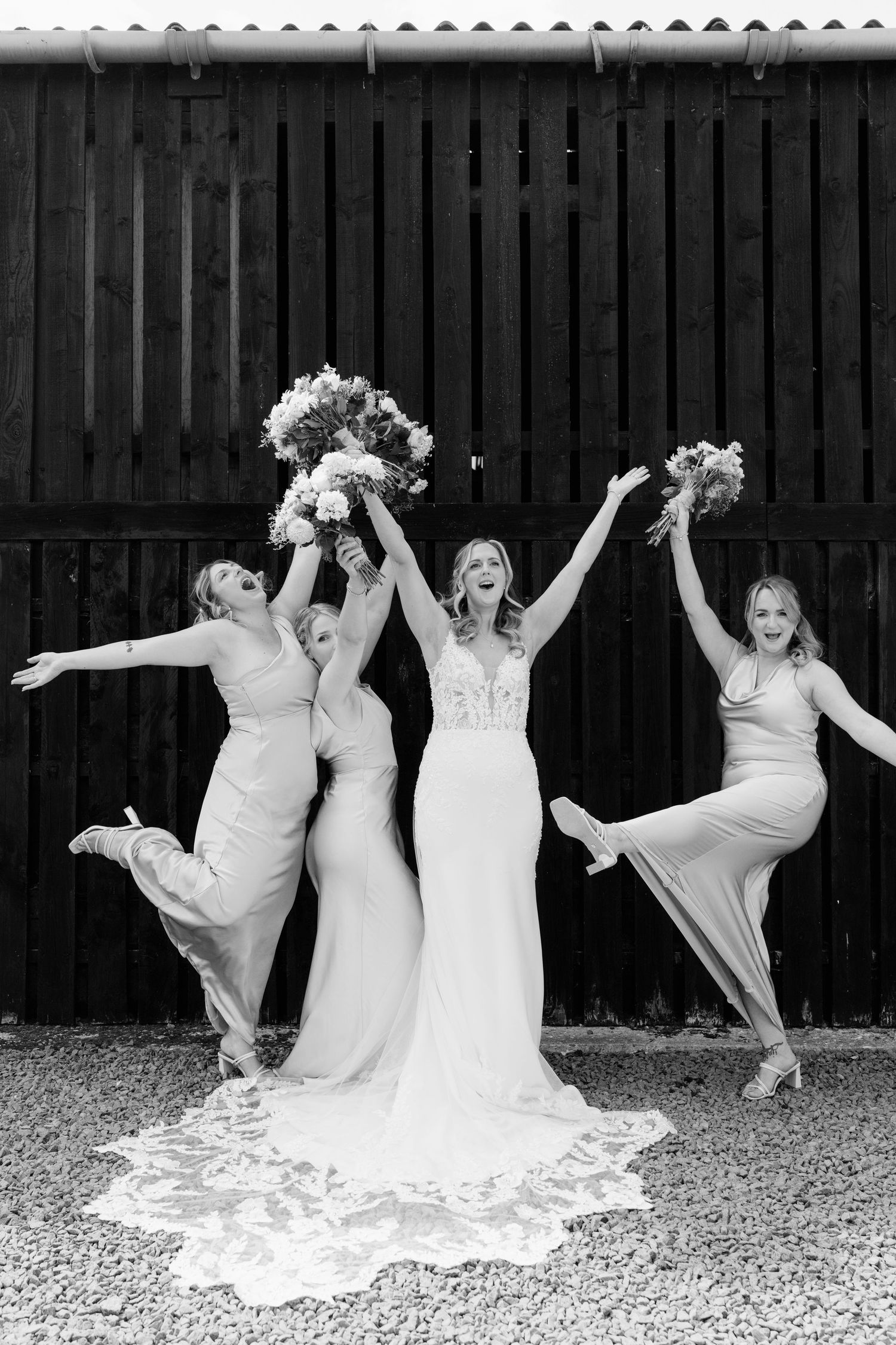 Black and white photo of a bride and three bridesmaids joyfully jumping and raising bouquets outside a barn.