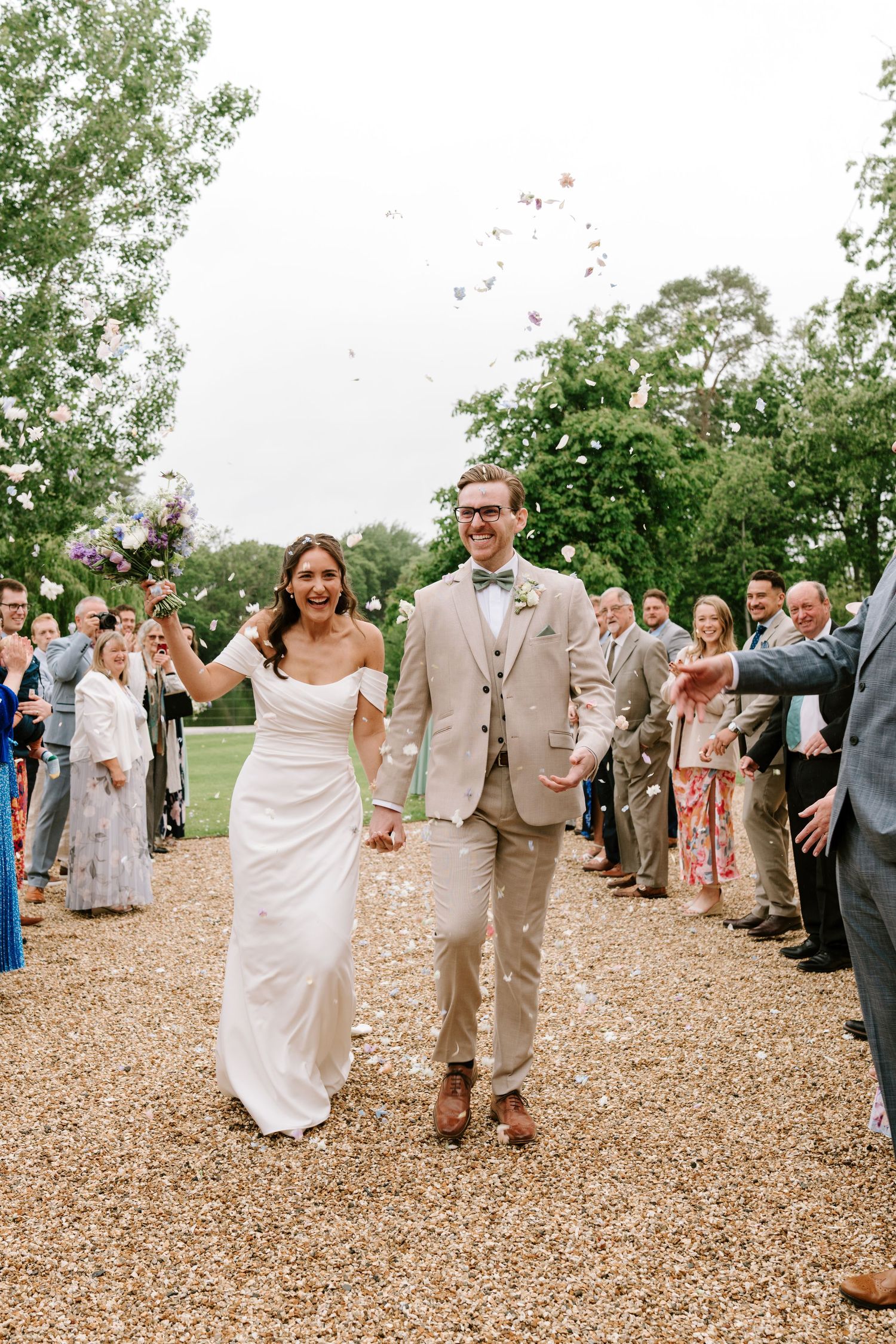Joyful bride and groom walk through colorful confetti toss on gravel path surrounded by cheering wedding guests.
