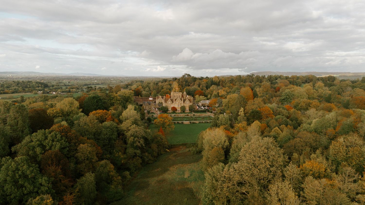 Aerial view of a grand historic mansion surrounded by autumn trees with golden foliage under a cloudy sky.