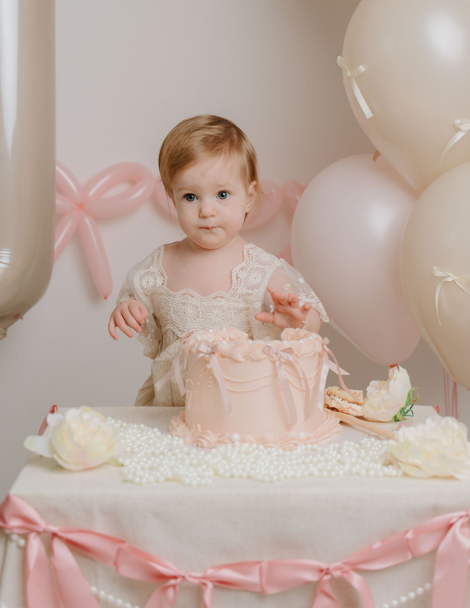 Baby girl in lace dress at first birthday cake smash with pink balloons and floral decorations.