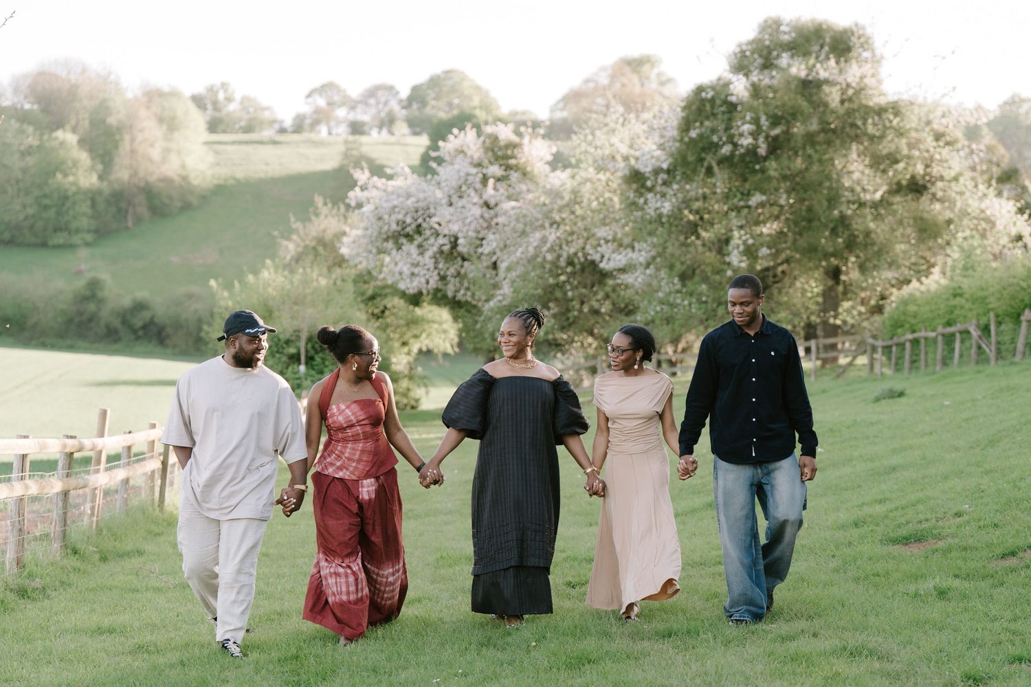 Five family members walk hand-in-hand across green hills with blooming trees in background.