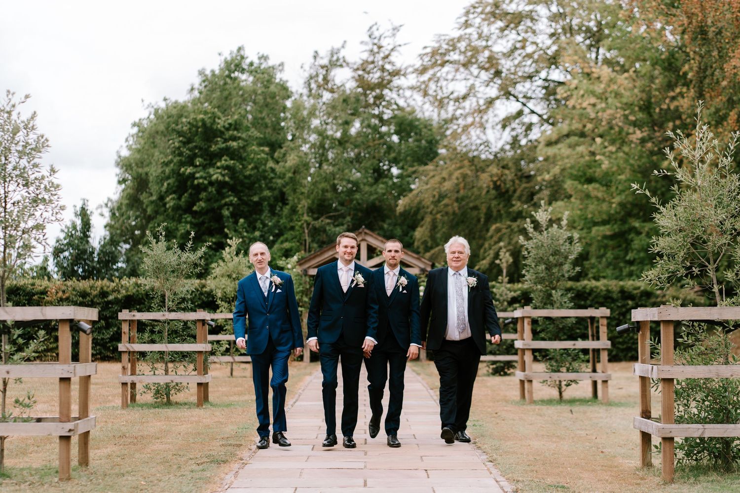 Four groomsmen in dark suits walking together along a garden path at an outdoor wedding venue.