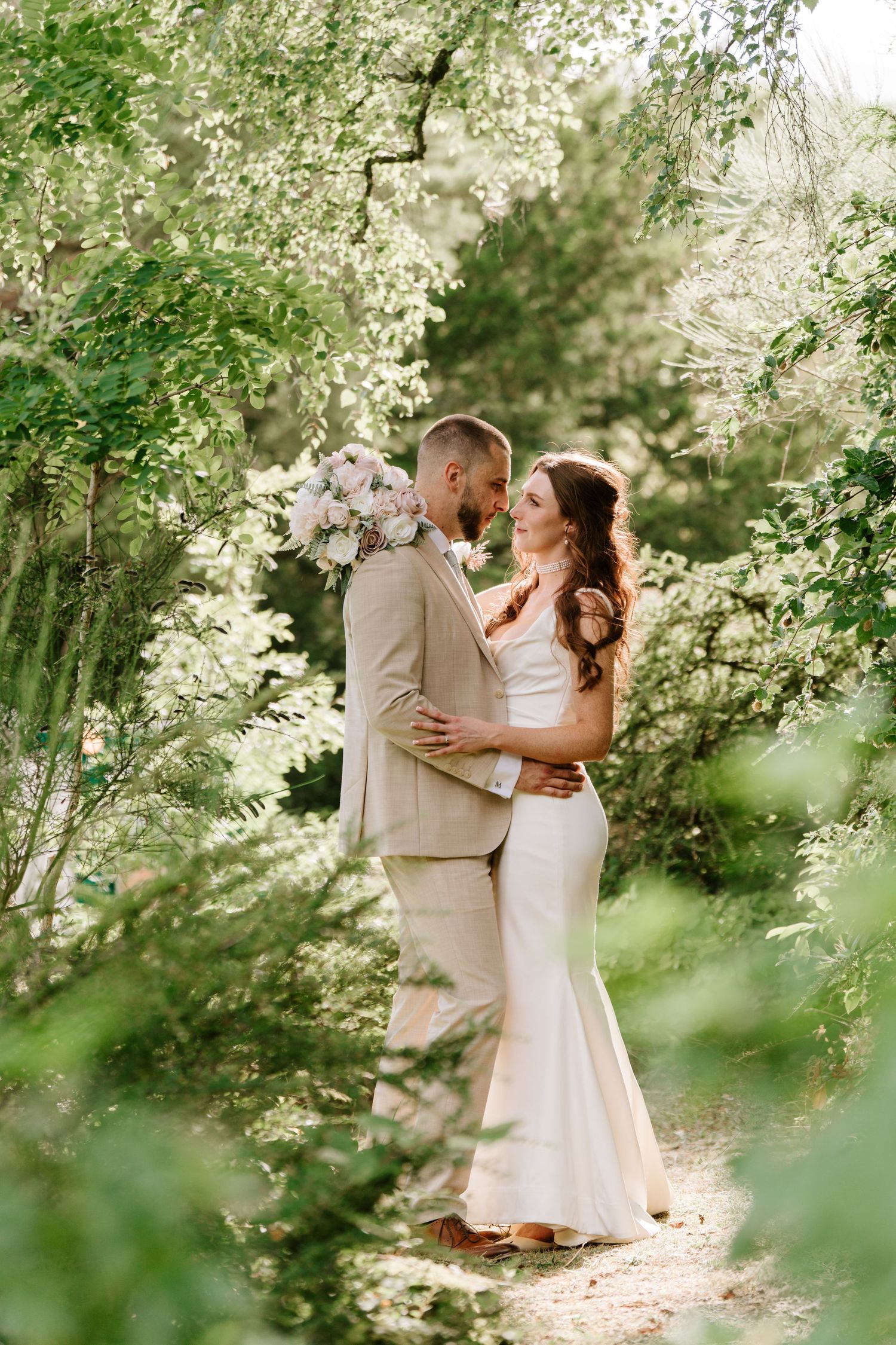 A bride and groom embrace in a lush green garden, wearing a white gown and tan suit, surrounded by blooming trees.