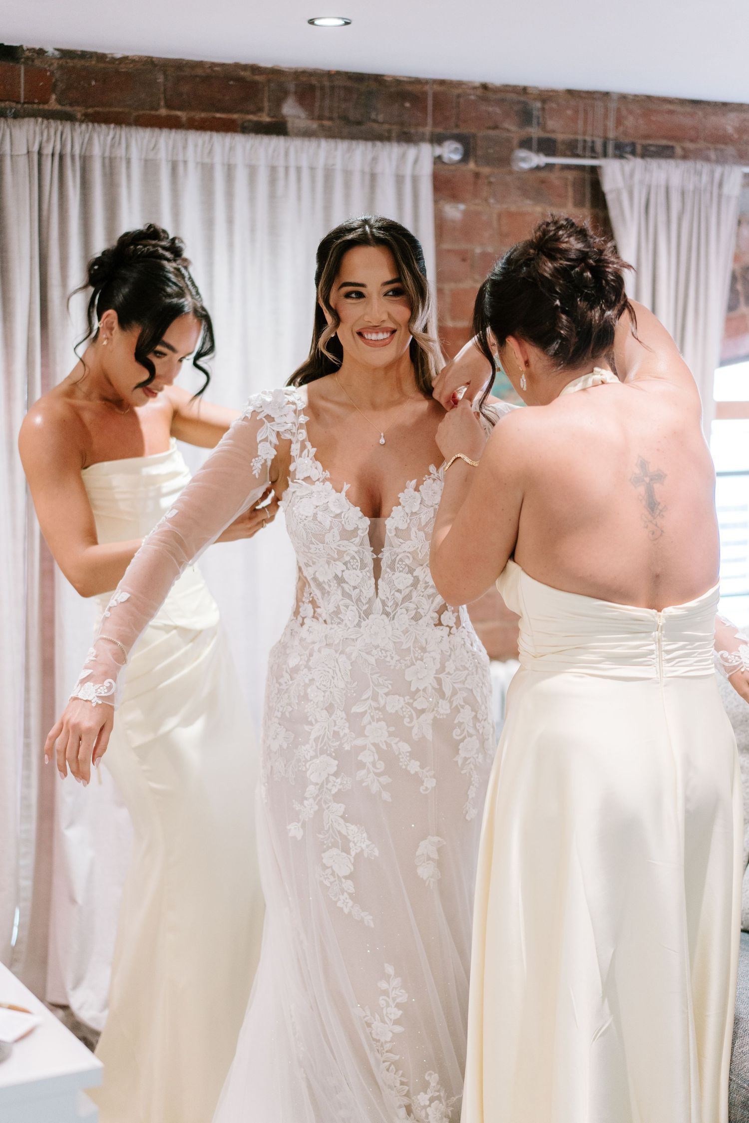Bride in lace wedding gown smiles as two bridesmaids in ivory dresses help her prepare for her wedding day.